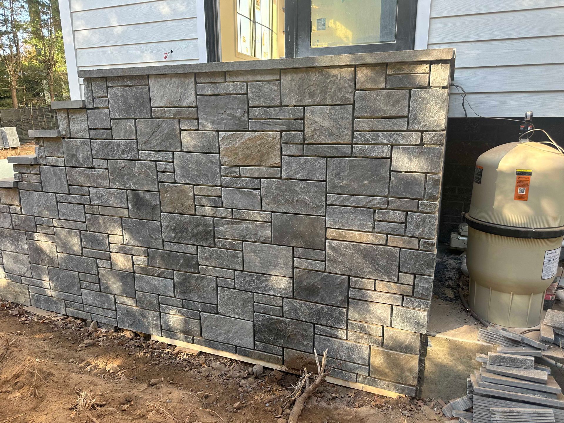 Stone veneer wall under construction next to a house with a pool filter.