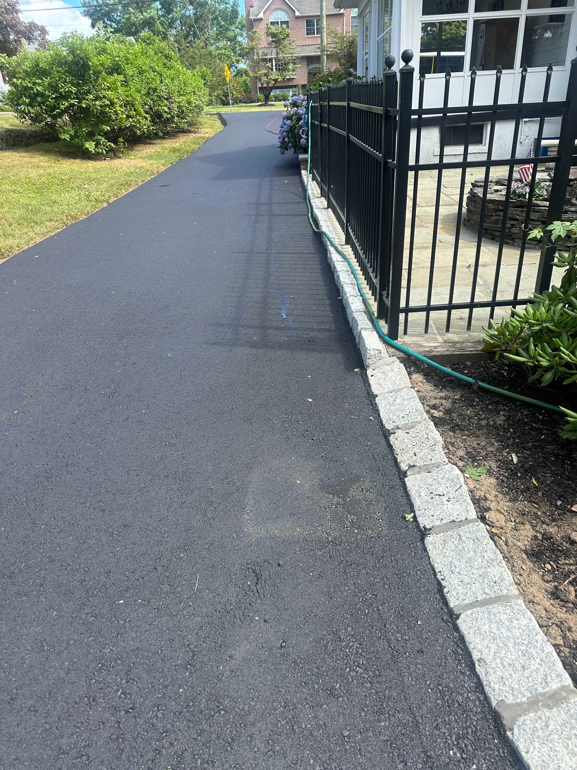 Black asphalt driveway with a black metal fence and stone border.