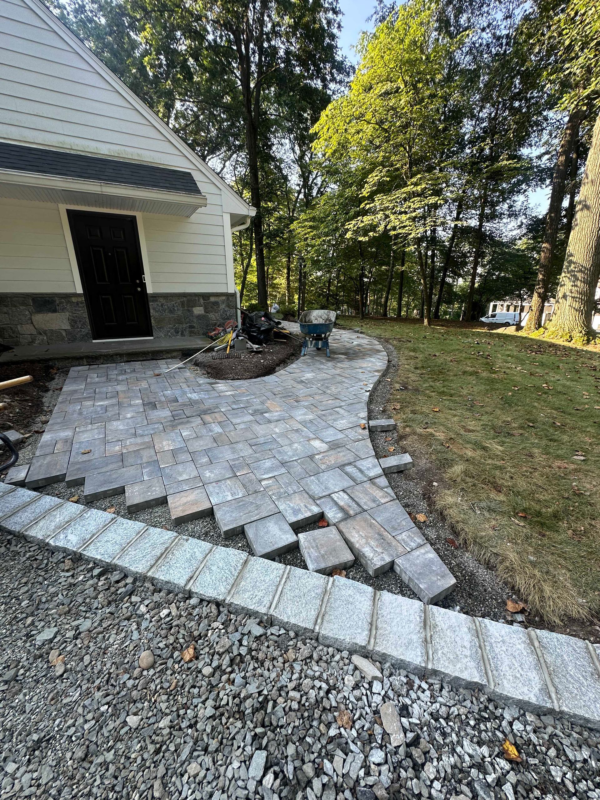 Stone patio and walkway being constructed alongside a light-colored building with dark door; green trees in the background.