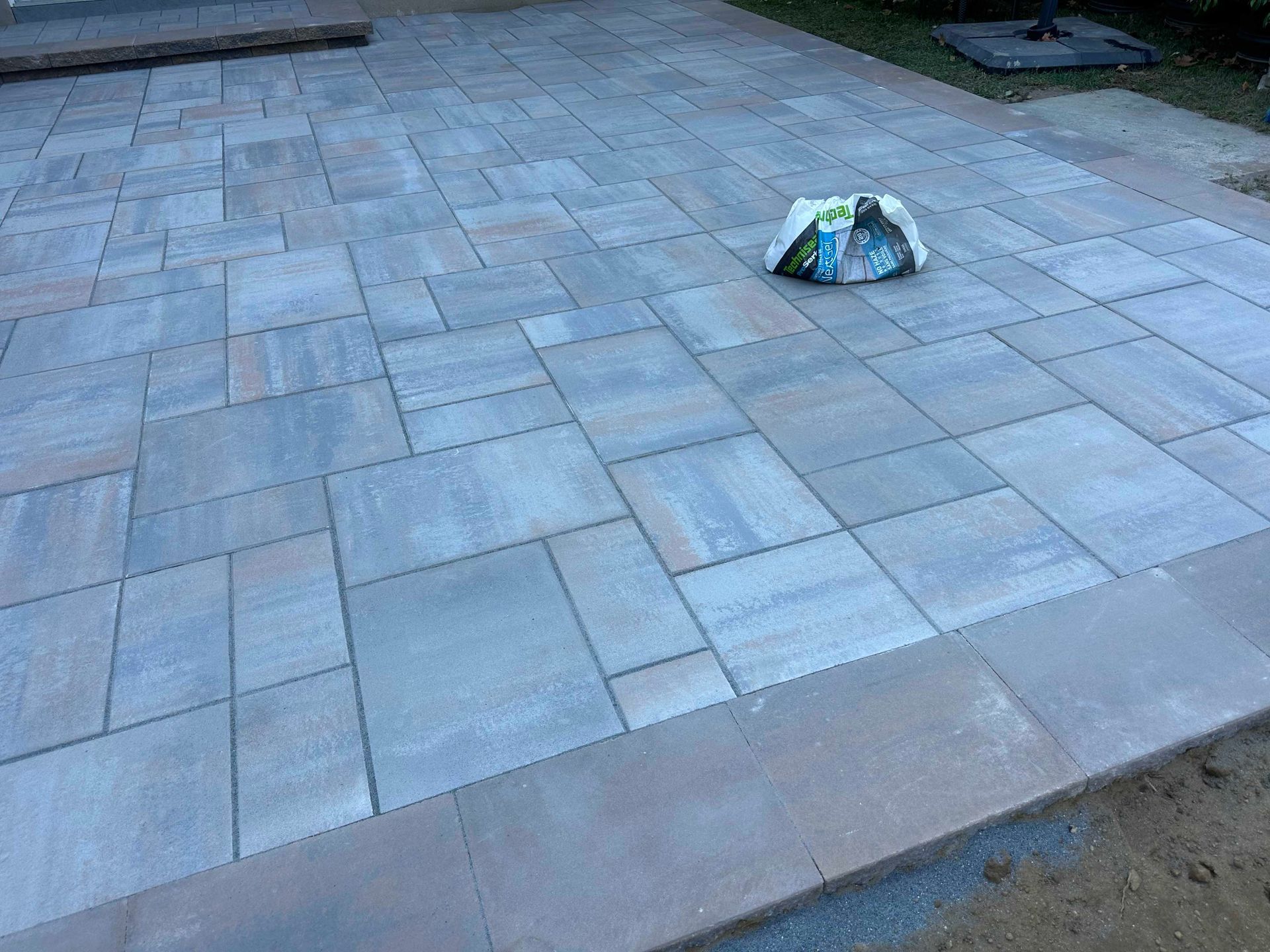 Paver patio with a bag on the surface. Gray and tan tones with a brown border.