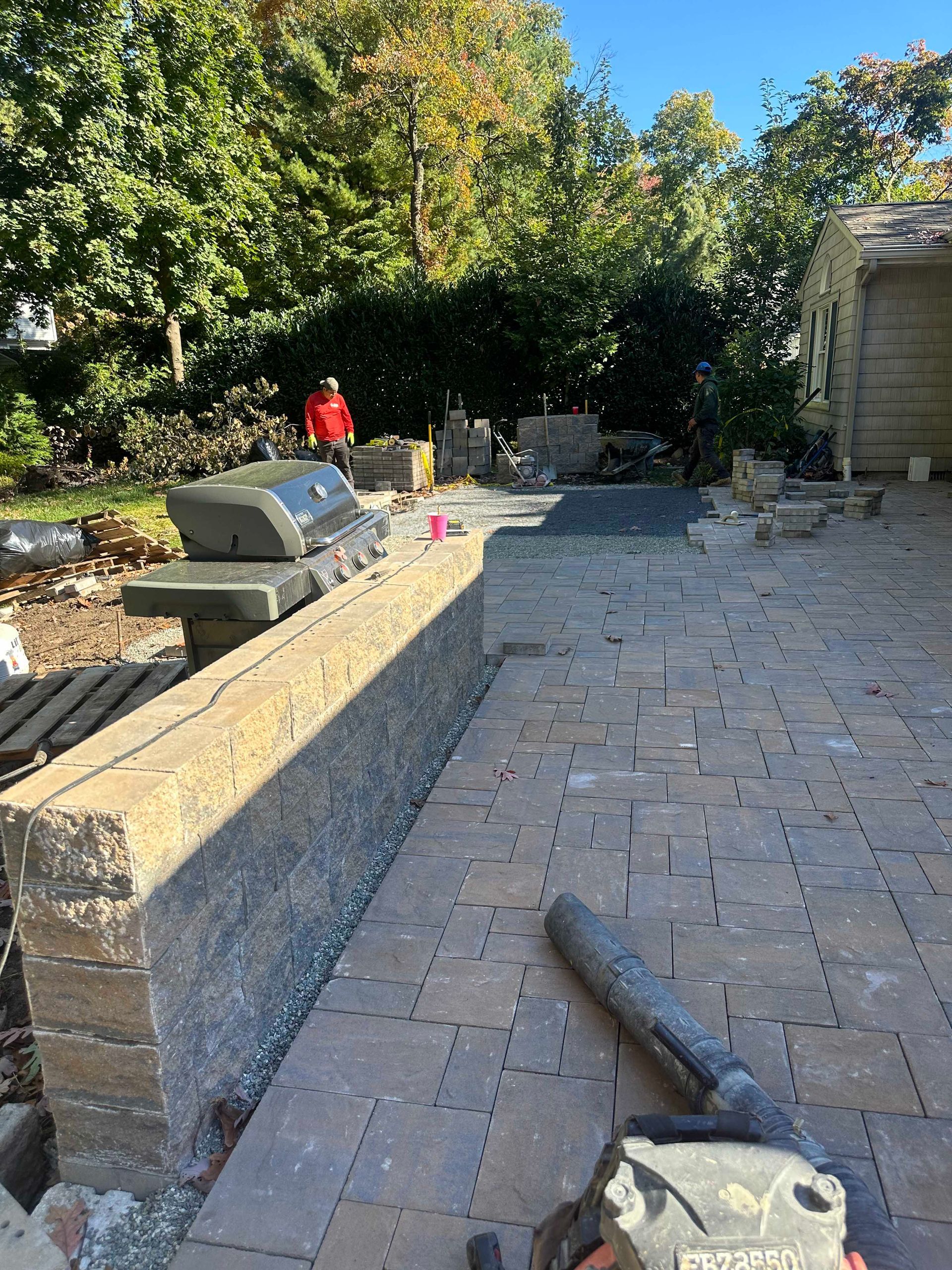 Patio construction with brick wall, grill, pavers, and blower. Person in red jacket in background.
