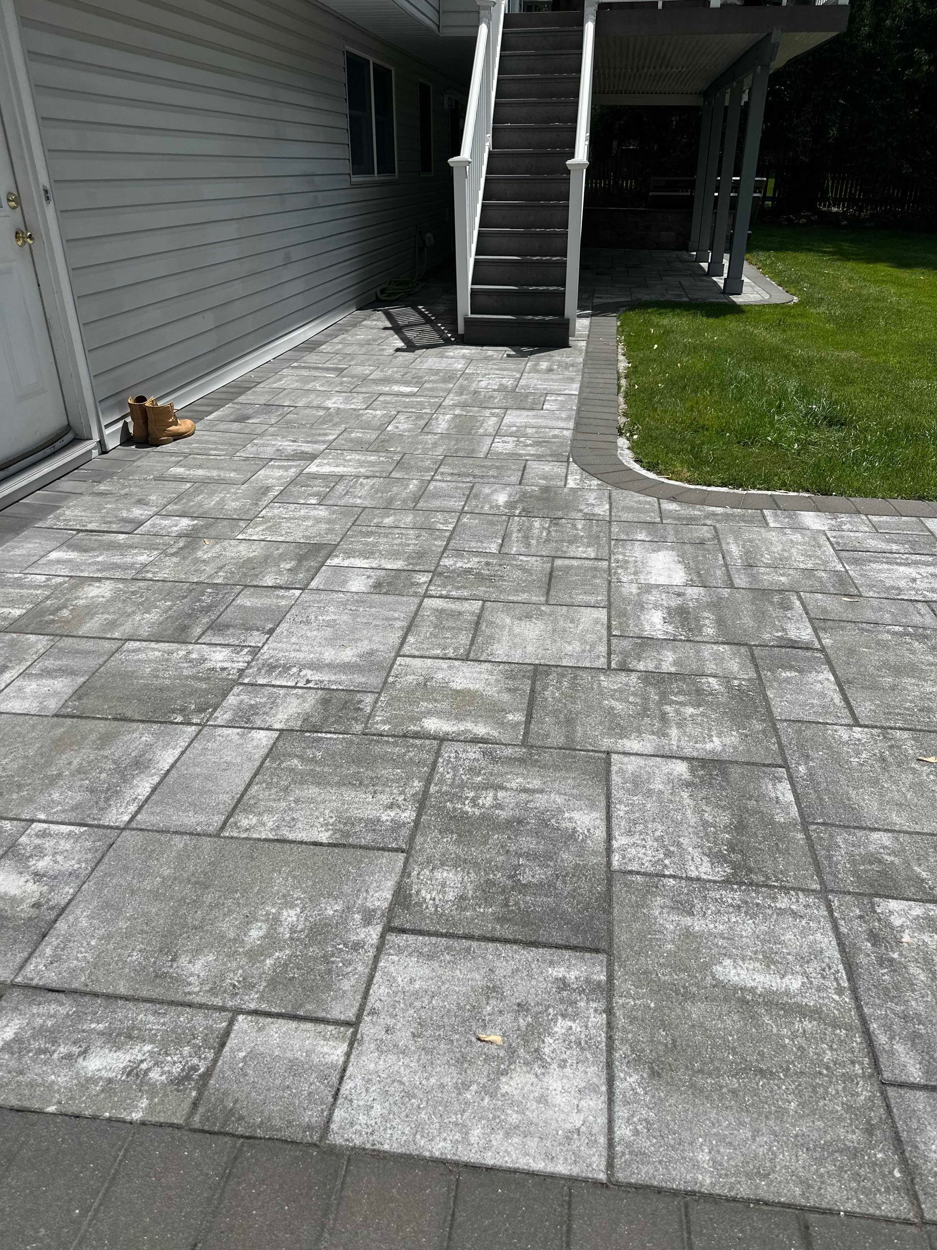 Gray patio pavers leading to a staircase, next to green grass and a house.