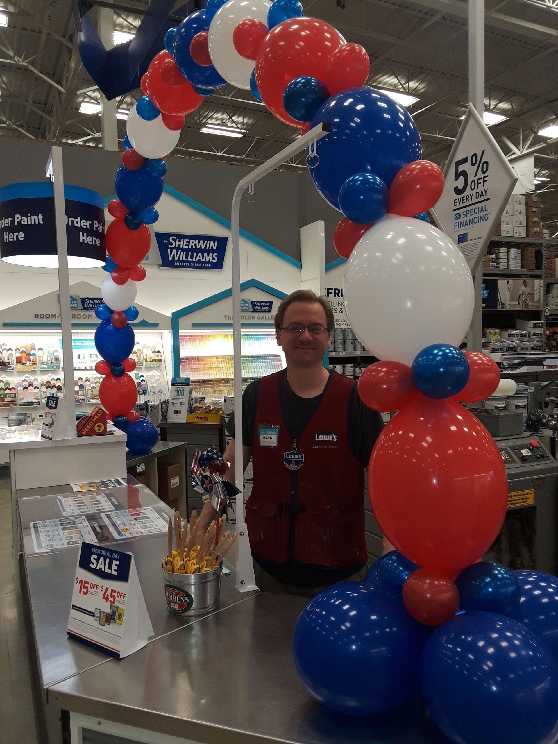 A man stands behind a counter decorated with red white and blue balloons