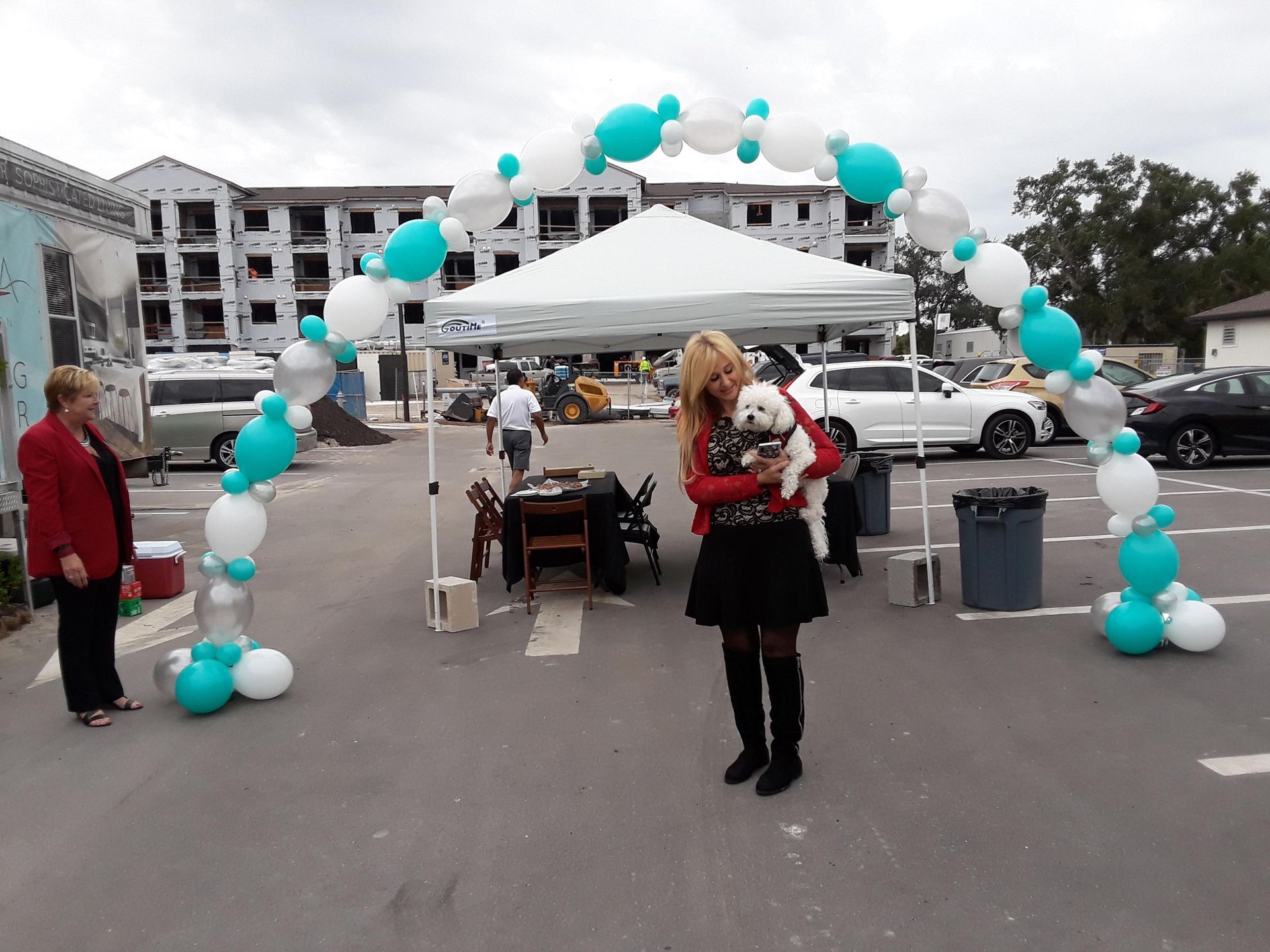 A woman is holding a small white dog in a parking lot surrounded by balloons.