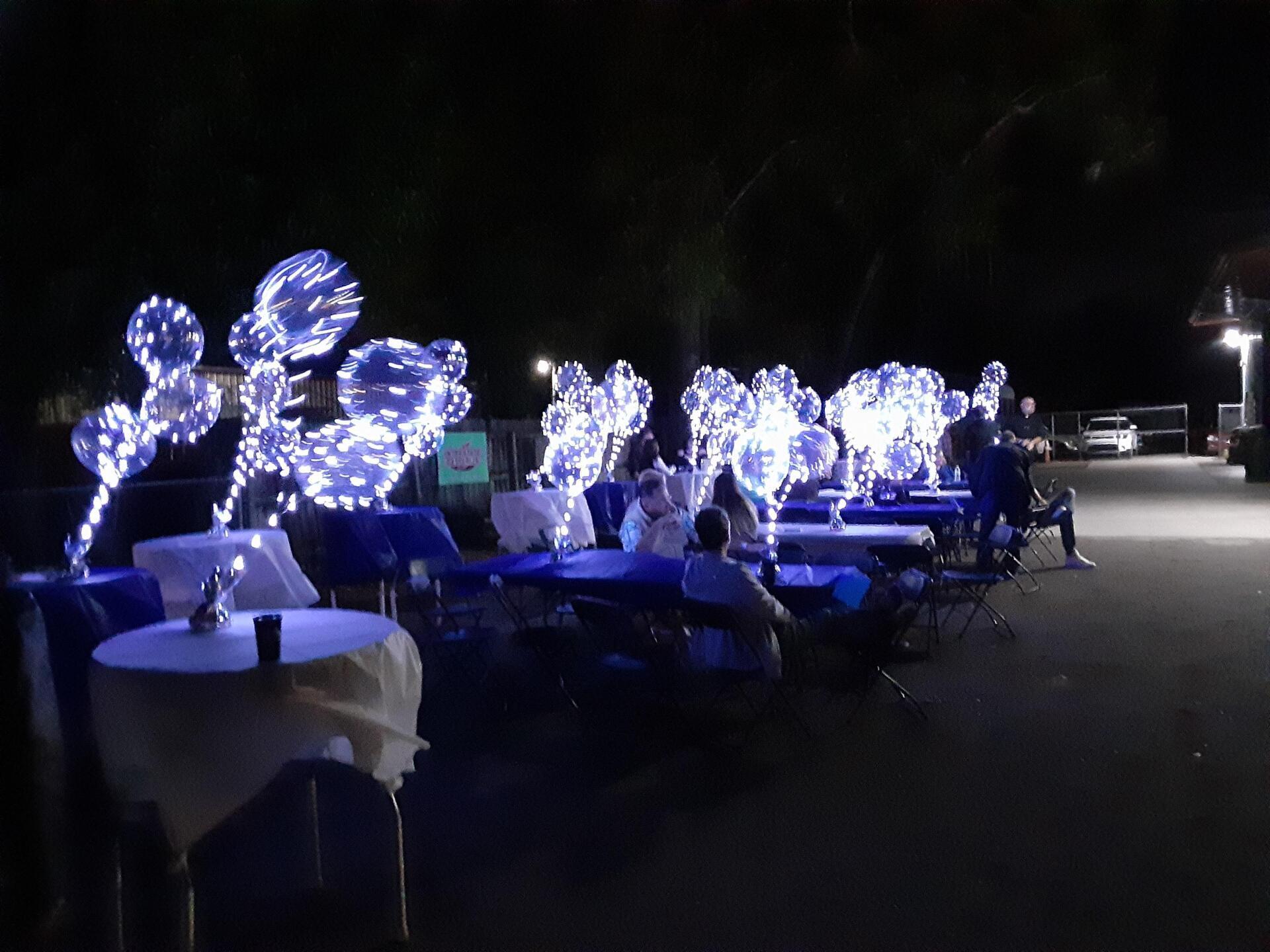 A group of people are sitting at tables in a parking lot at night.