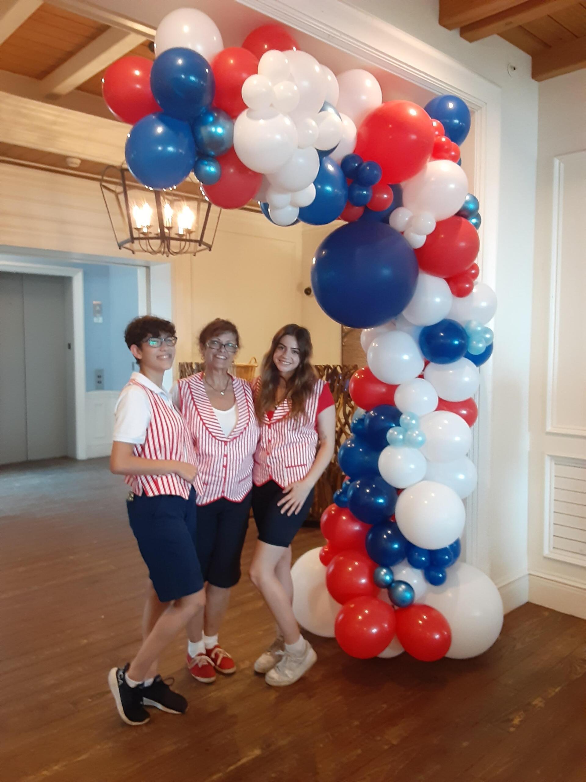 Three people standing in front of a red white and blue balloon arch