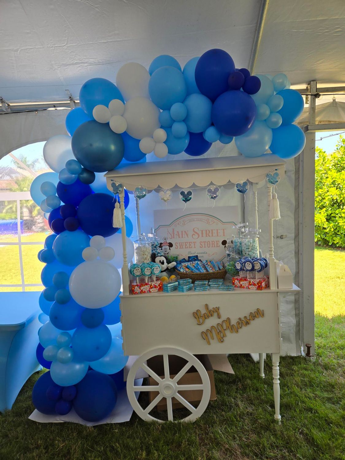 A candy cart is decorated with blue and white balloons for a baby shower. 