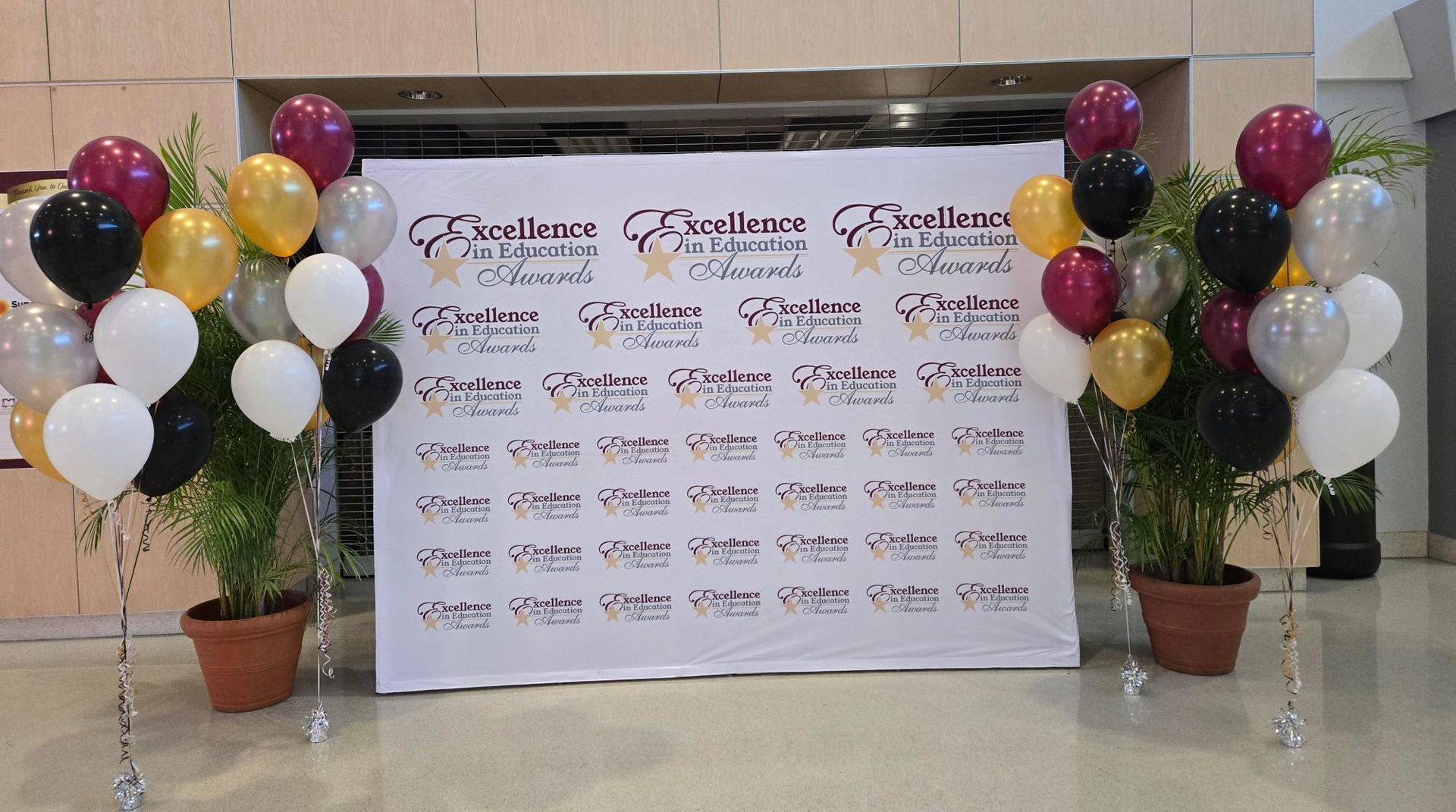 Balloons in various colors flank a white banner with text and logos, flanked by potted plants, likely for an event.