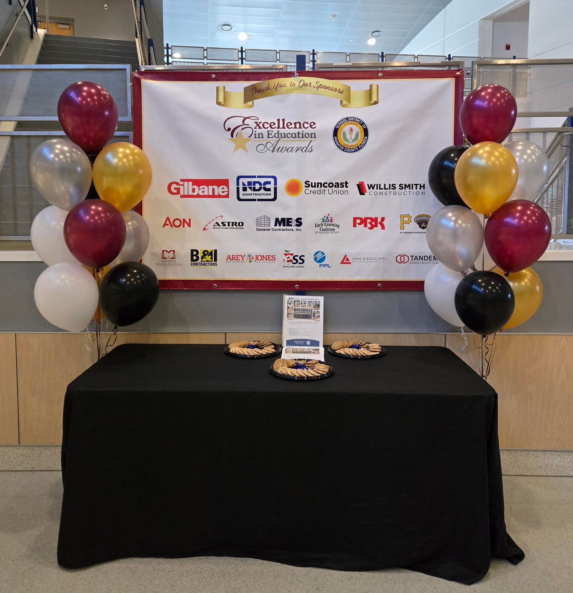 Table with banner for an event, balloons on each side, black tablecloth, gold, black, silver, burgundy, and white balloons.