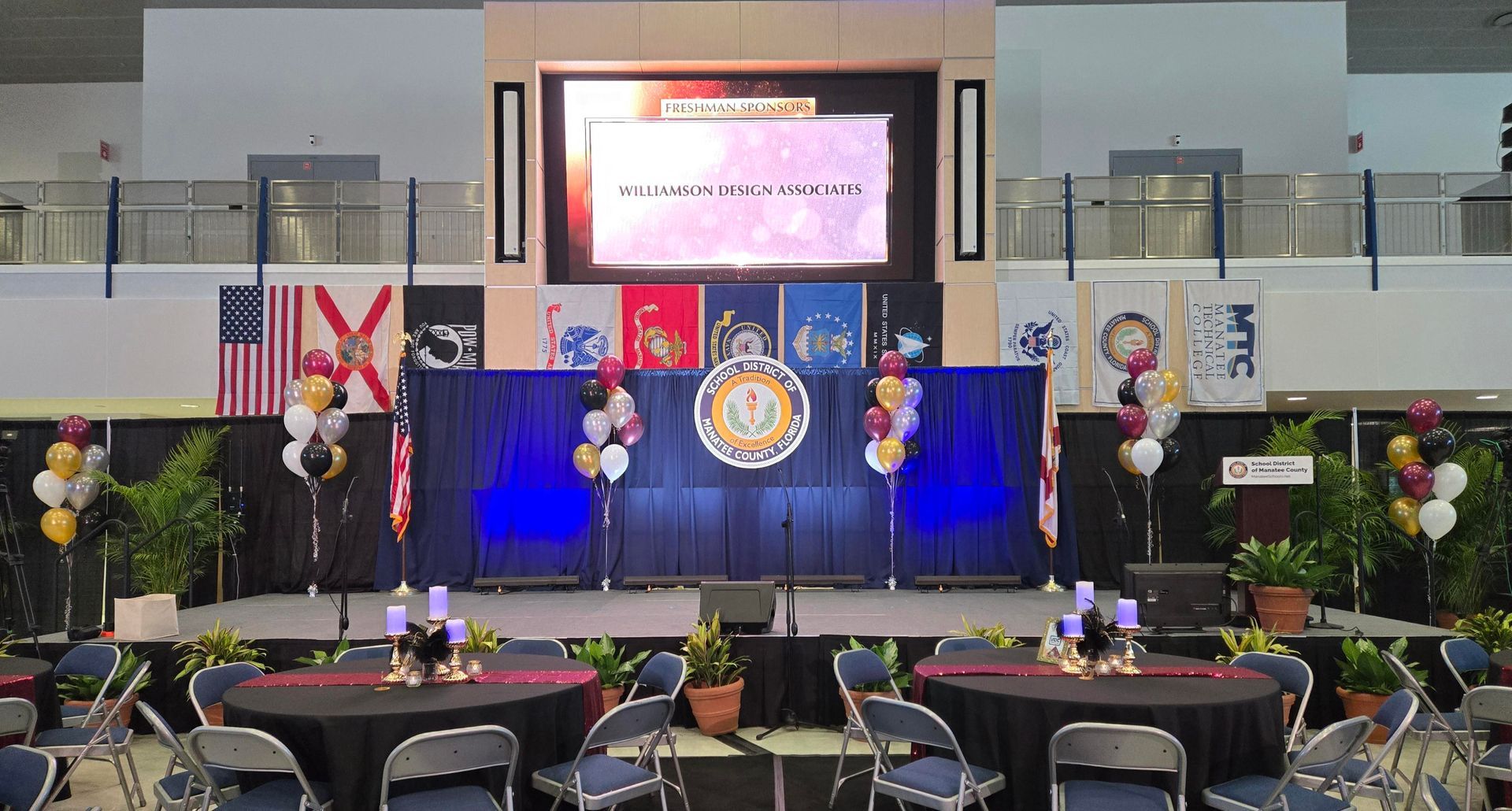 Stage setup with banners, balloons, tables, and a large screen for an event.