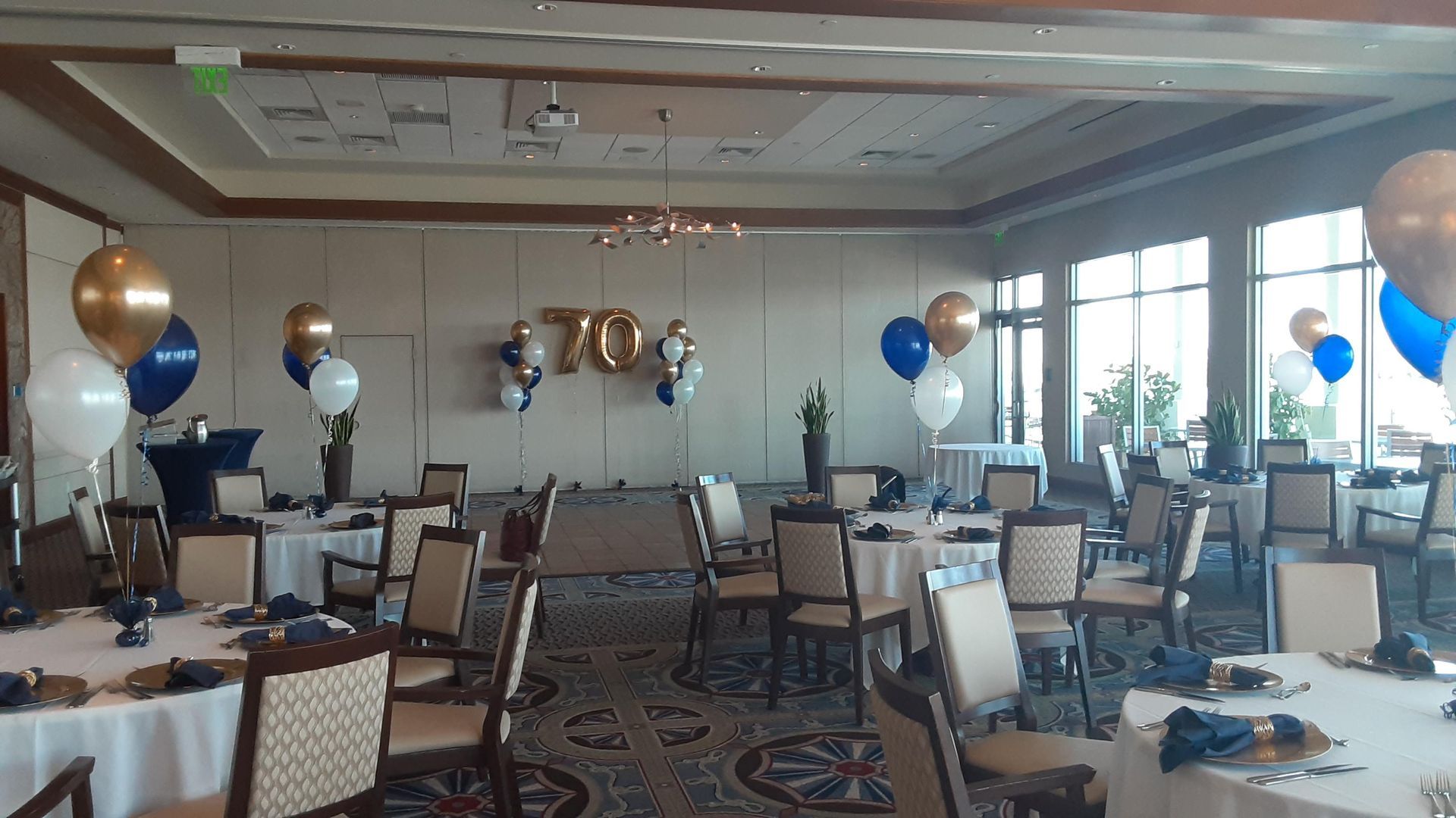 Room set up for a 70th birthday celebration. Round tables with white linens, blue and gold balloons.