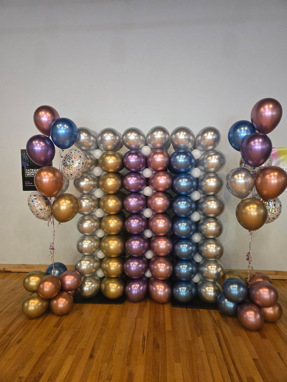 Balloon backdrop with metallic hues, flanked by balloon clusters and bouquets, set on wooden floor.