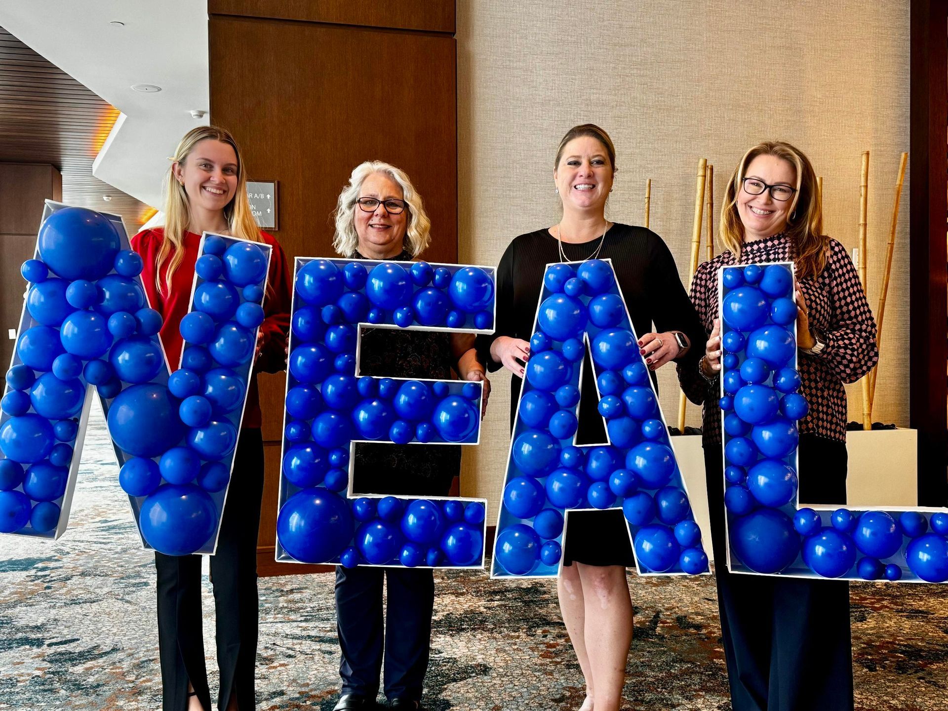 Four women holding a blue balloon-filled sign that spells NEAL, inside a building with a blurred background.