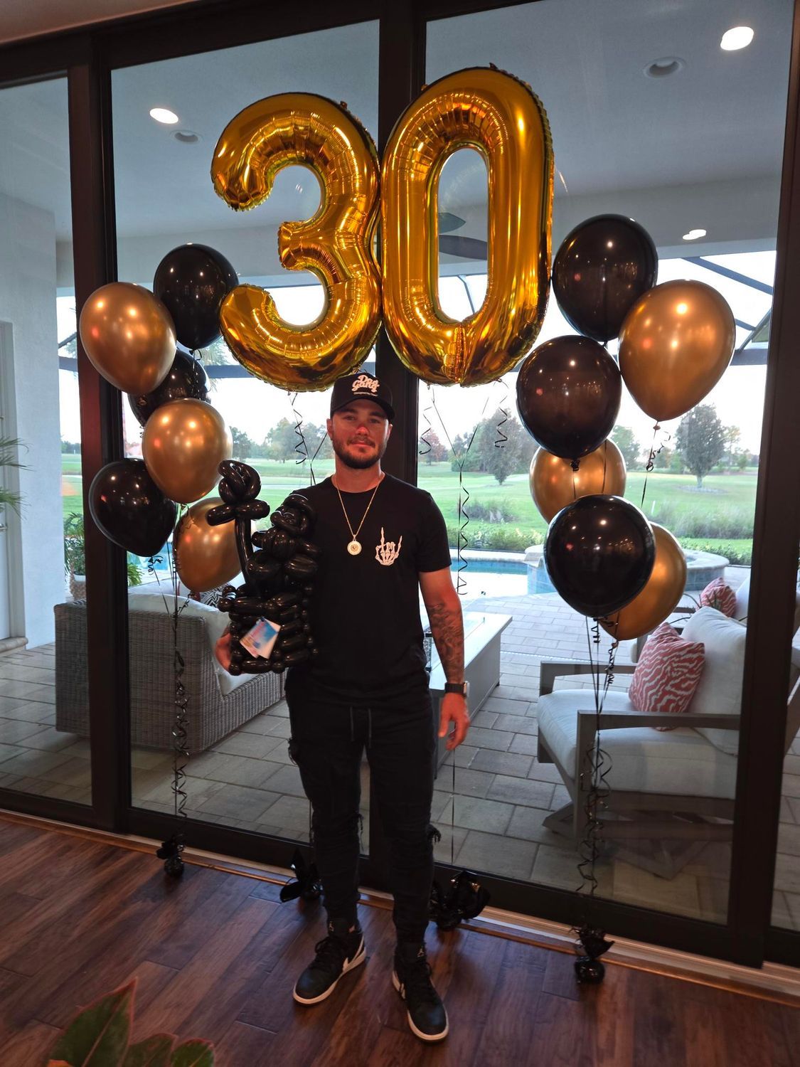 Man celebrating 30th birthday with gold and black balloons. He holds a smaller balloon bouquet; setting is a home with a pool.