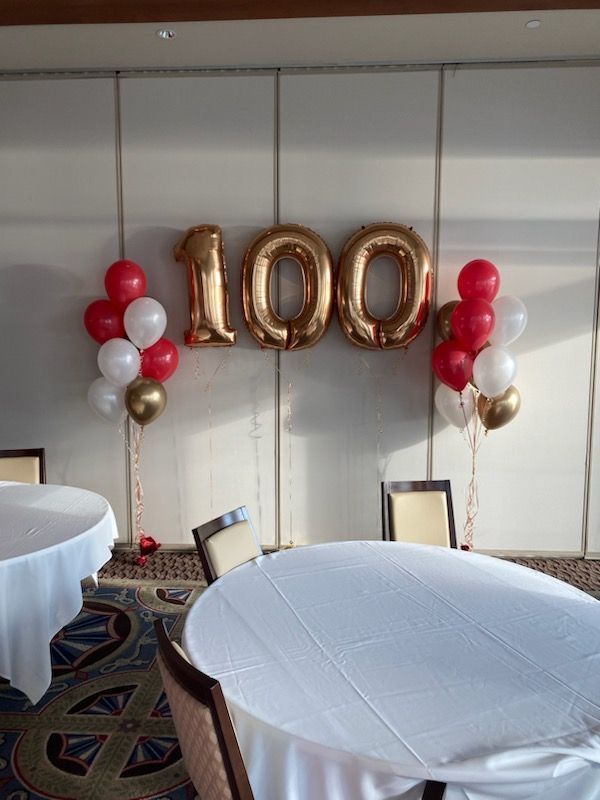Golden 100 balloons with red, white, and gold balloon bouquets, set against a white wall, celebrating a milestone.
