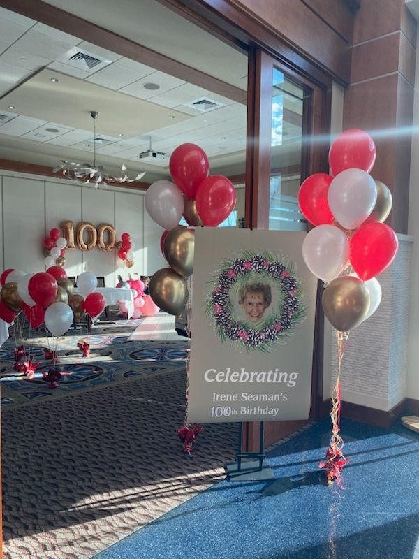 A 100th birthday celebration with red, white, and gold balloons, a sign with a photo, and the number 100 in gold balloons.