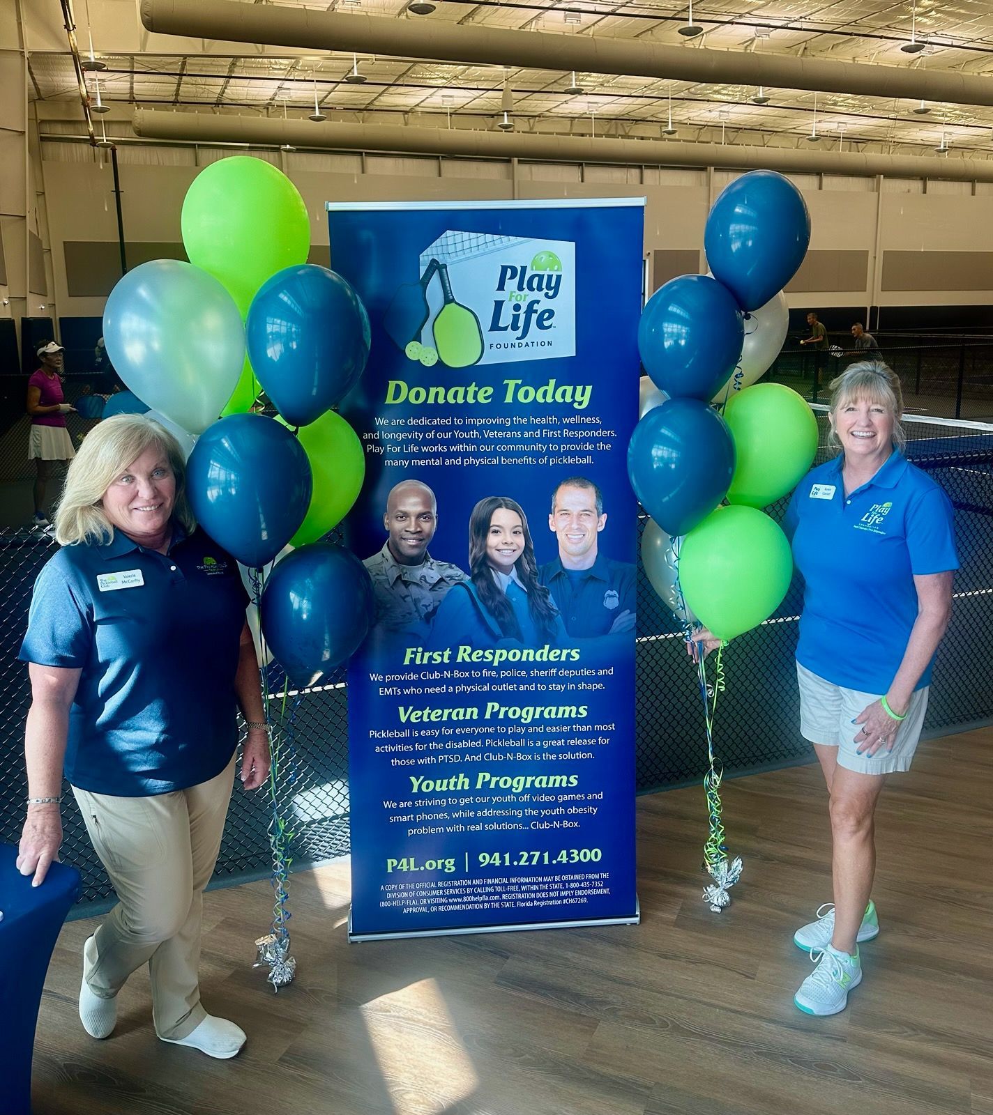 Two women standing by a Play Life banner with balloons, promoting donations for veterans and first responders.