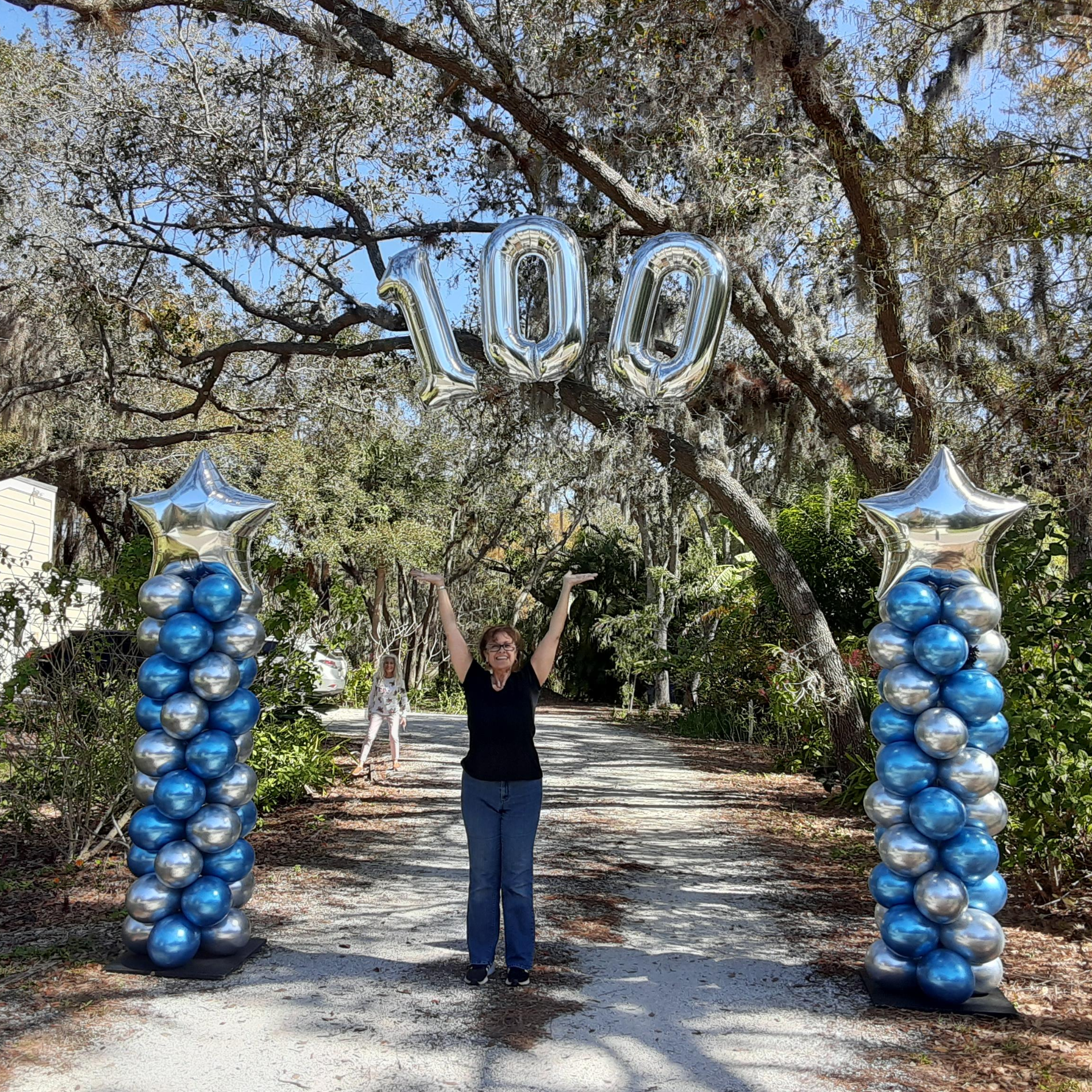Woman celebrates a 100th birthday with balloons under a tree archway, with two columns of blue/silver balloons.