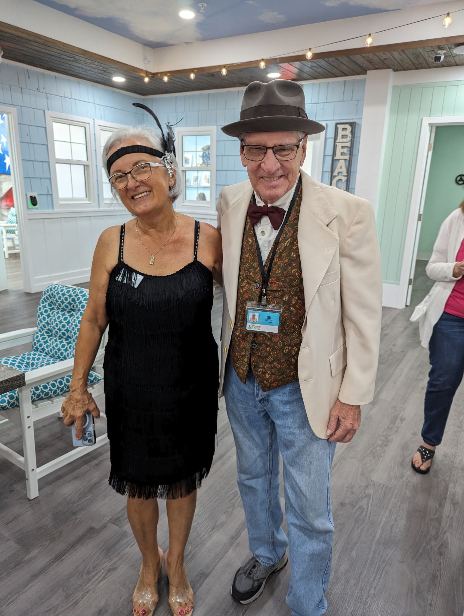 Couple dressed in 1920s attire posing together indoors, smiling. The woman in a black dress, the man in a suit and hat.
