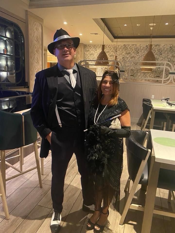 Couple in 1920s attire. Man in pinstripe suit, fedora; woman in flapper dress, boa. Inside restaurant.