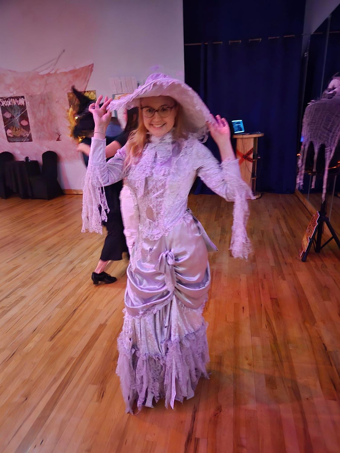 Woman in a lavender Victorian costume smiles, dancing on a wooden floor. She wears a large hat.