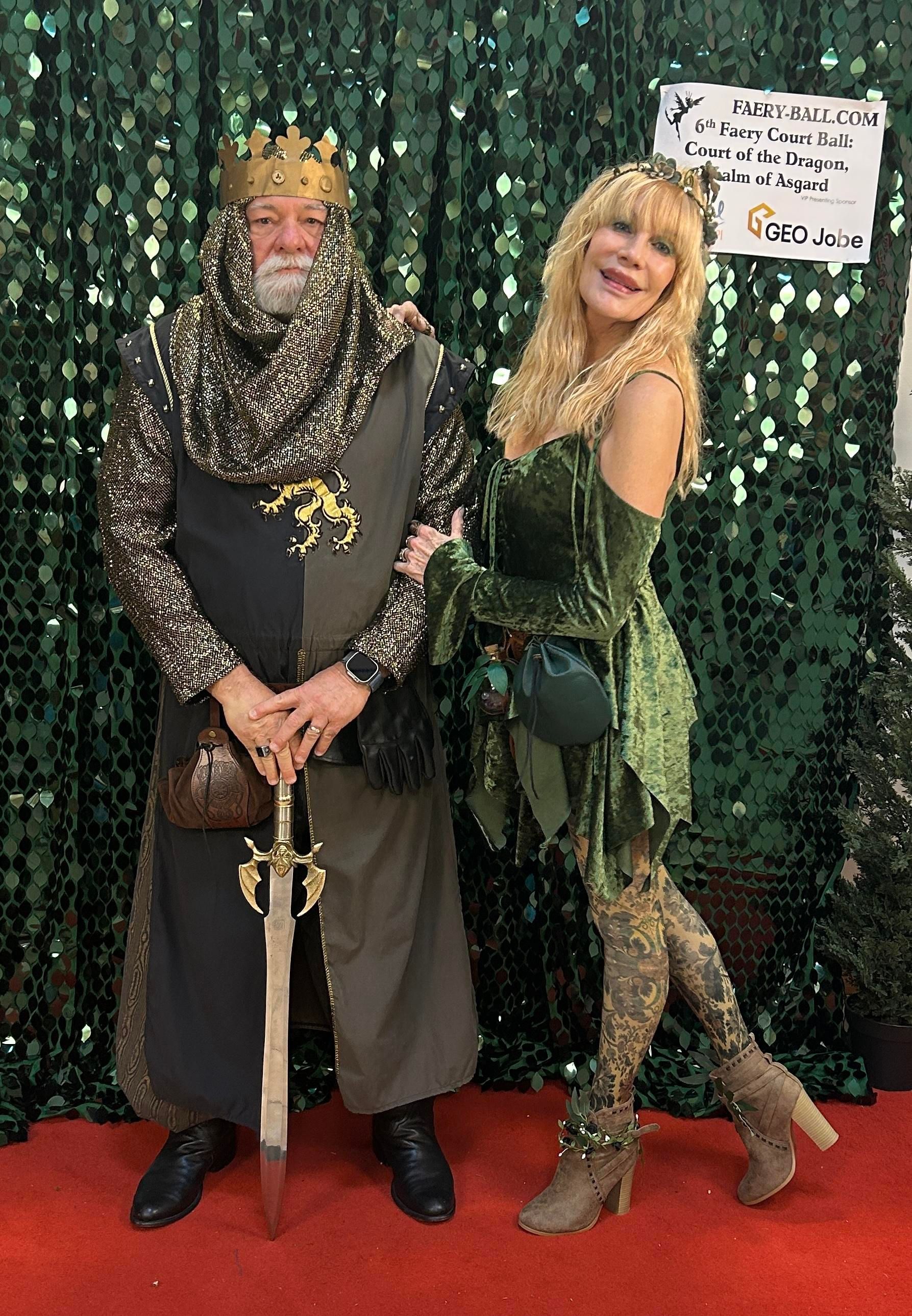 Man in king costume with a sword and woman in fairy costume pose in front of a green backdrop.