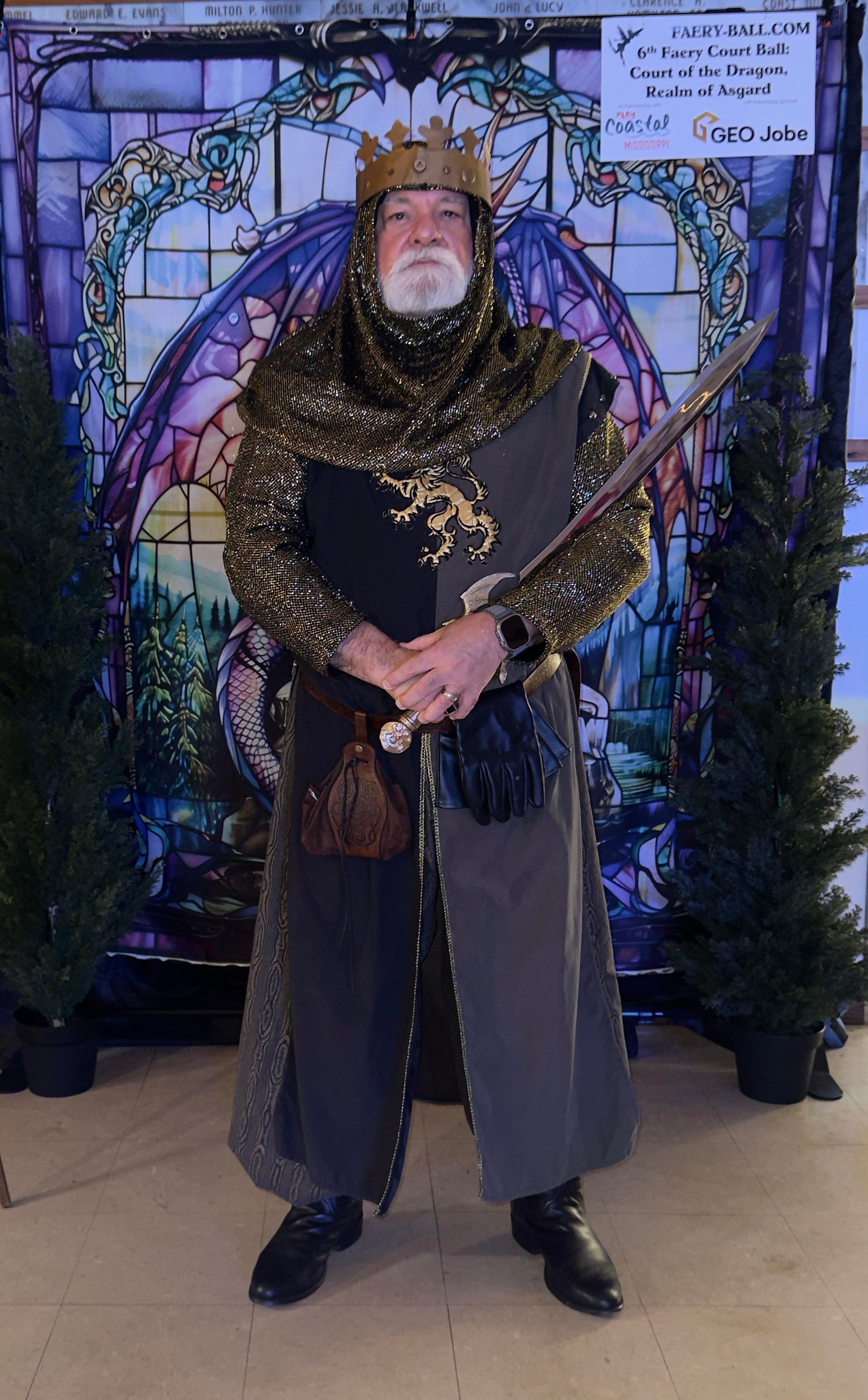 King in medieval attire, holding sword, stands before stained glass window.