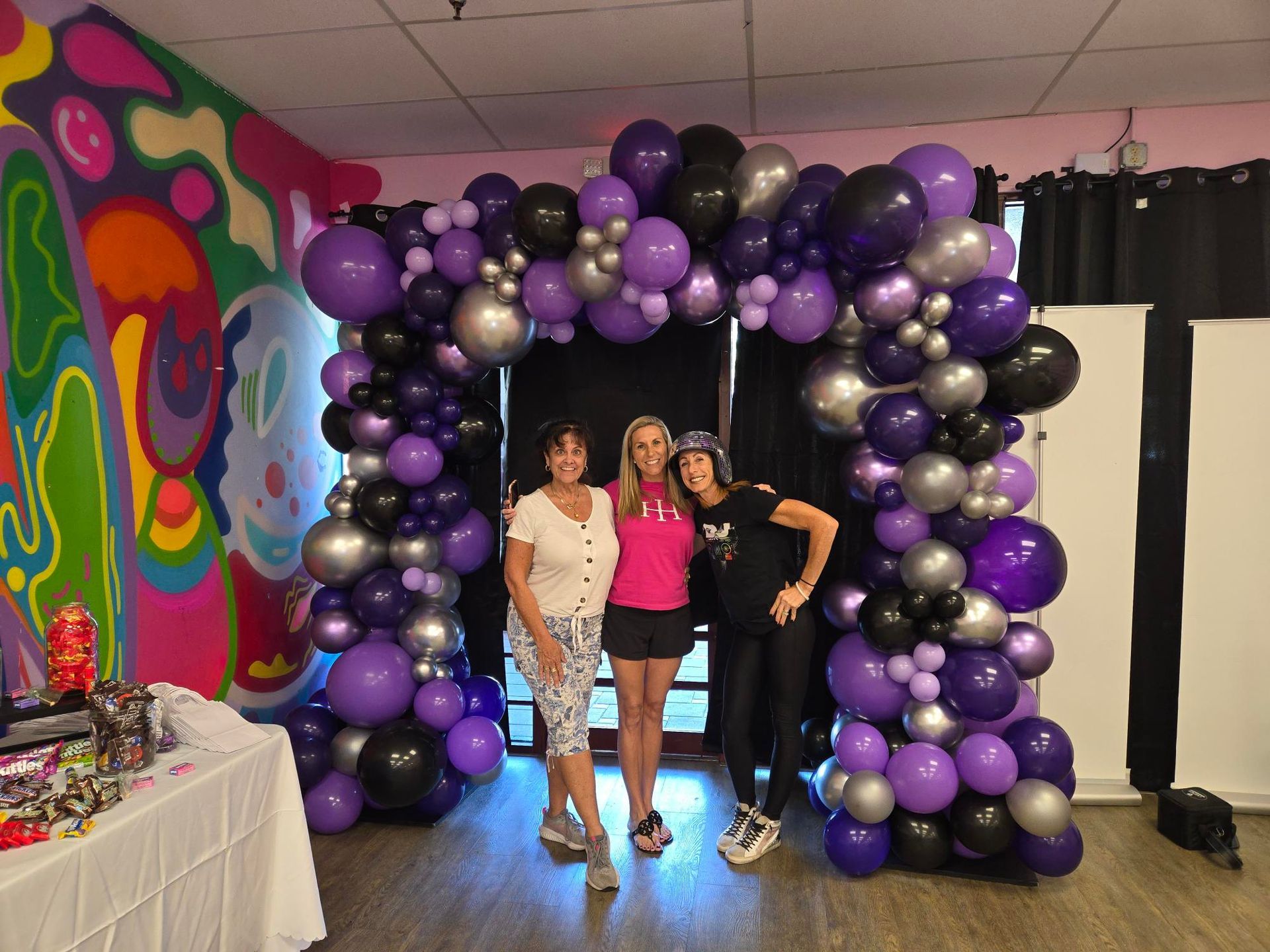 Three women pose under a purple, black, and silver balloon archway