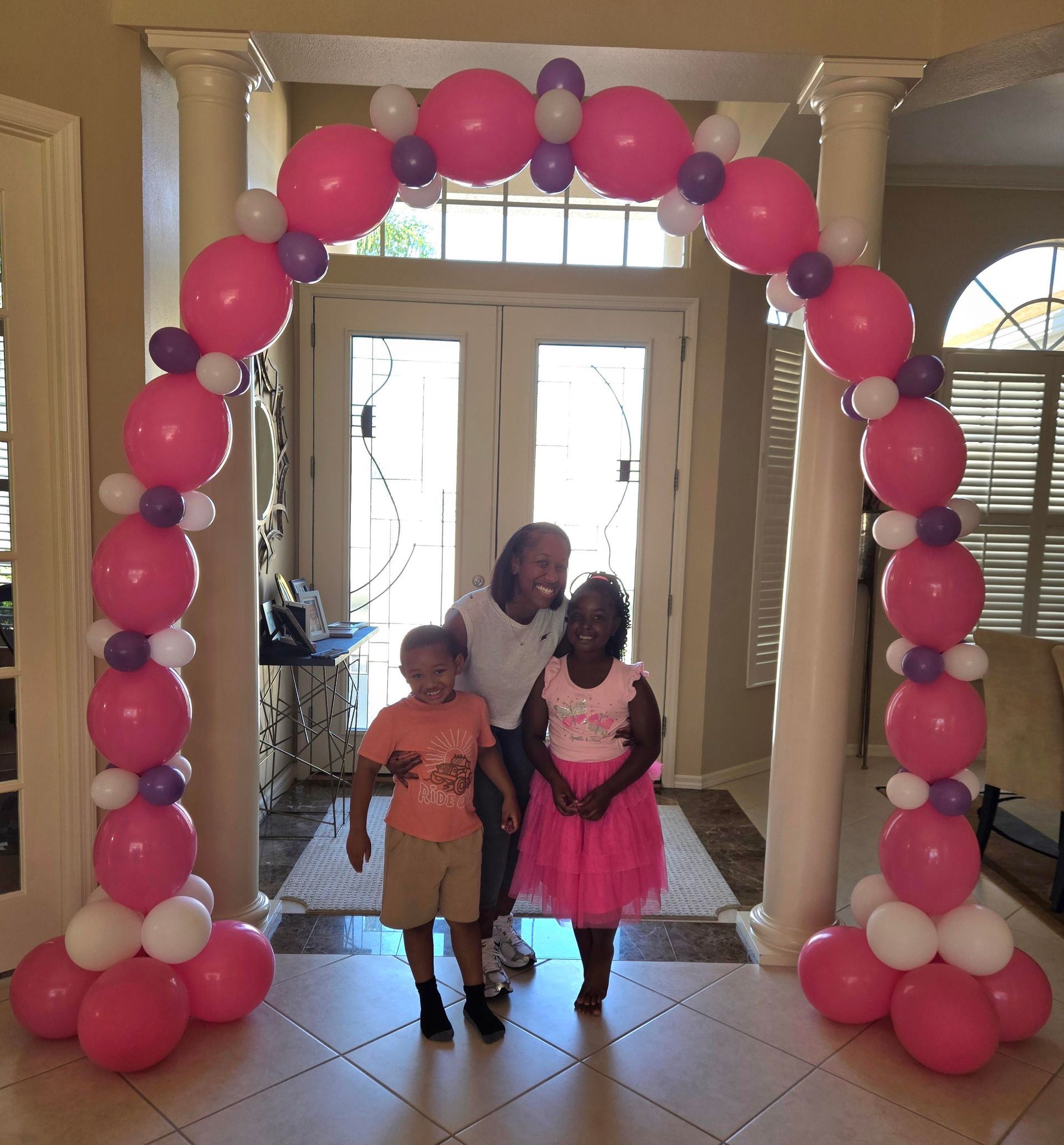 Children and a woman pose under a pink, white, and purple balloon arch in a doorway