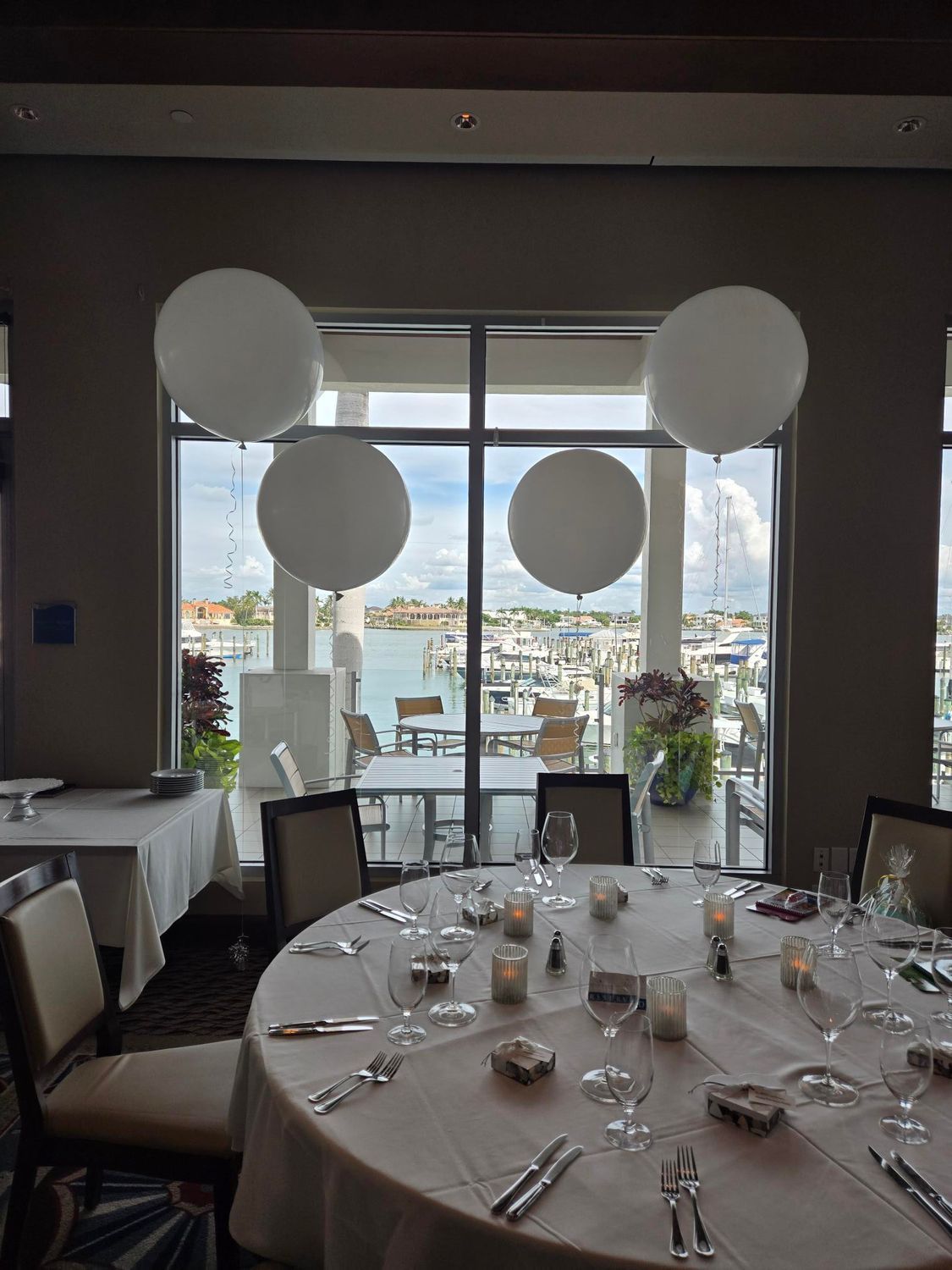 Four white balloons above a round table set for a formal event, with a harbor view through a window