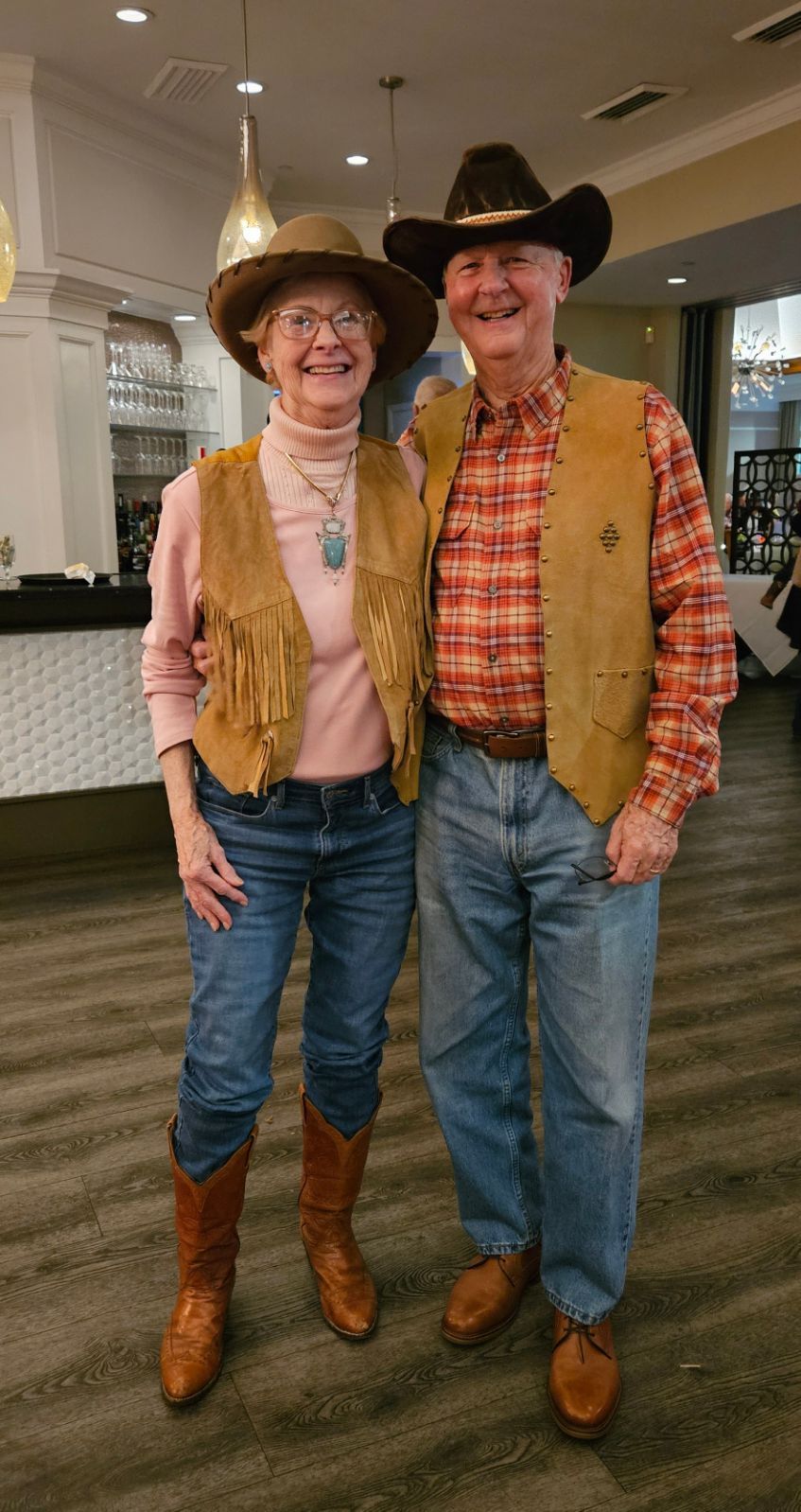Couple in cowboy attire, smiling. Woman wears a pink top, vest, jeans, boots