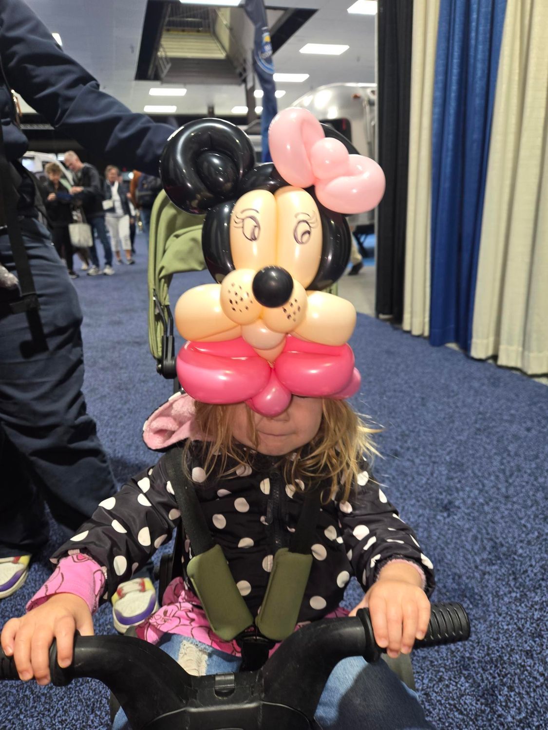 Child on a tricycle wearing a Minnie Mouse balloon hat. Indoors, blue carpet, polka dot jacket, smiling.