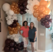 Two women pose beneath a balloon arch in brown, gold, white, and orange.