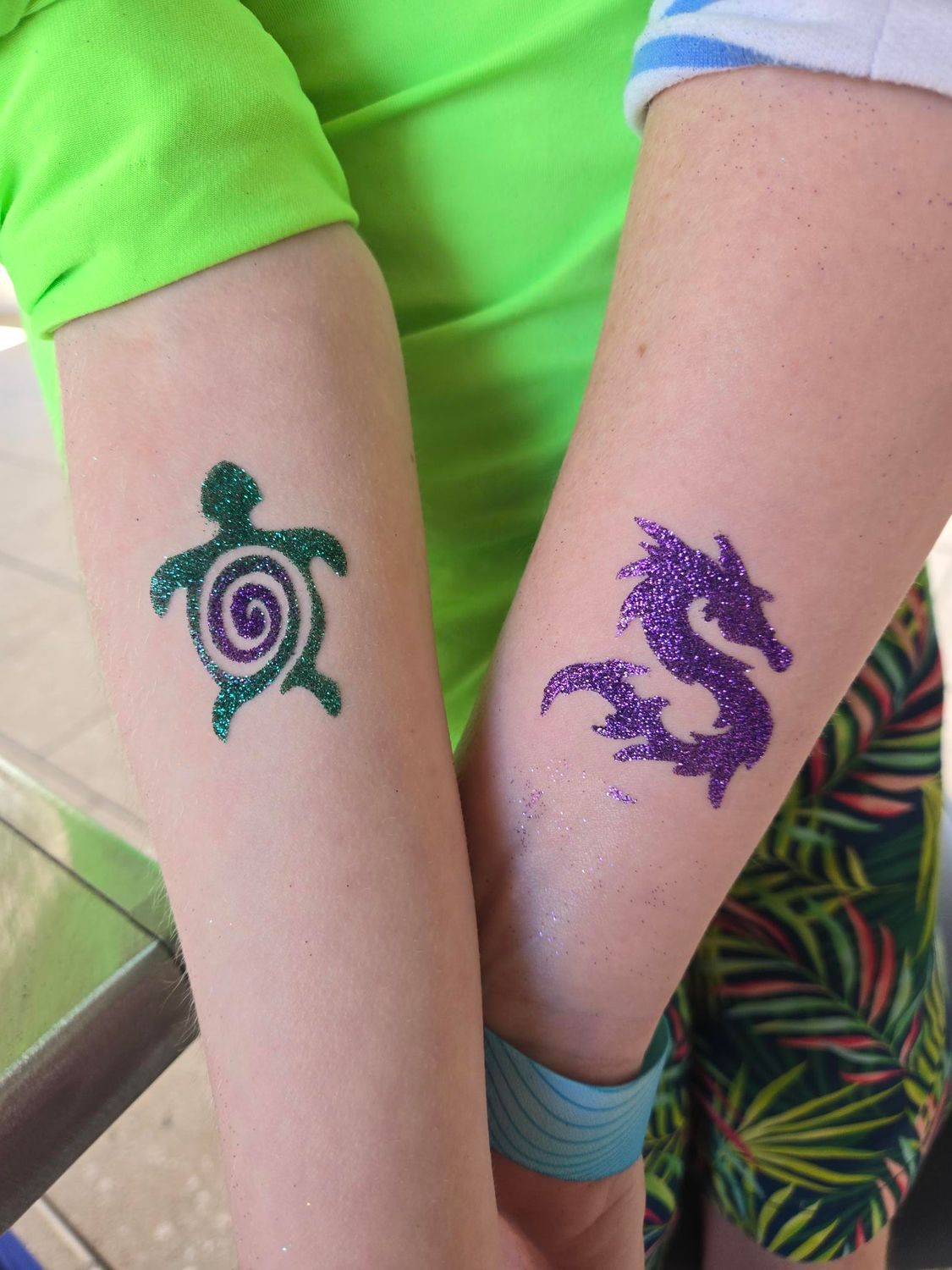 Two arms side-by-side, with glitter tattoos: turtle (green) and dragon (purple).