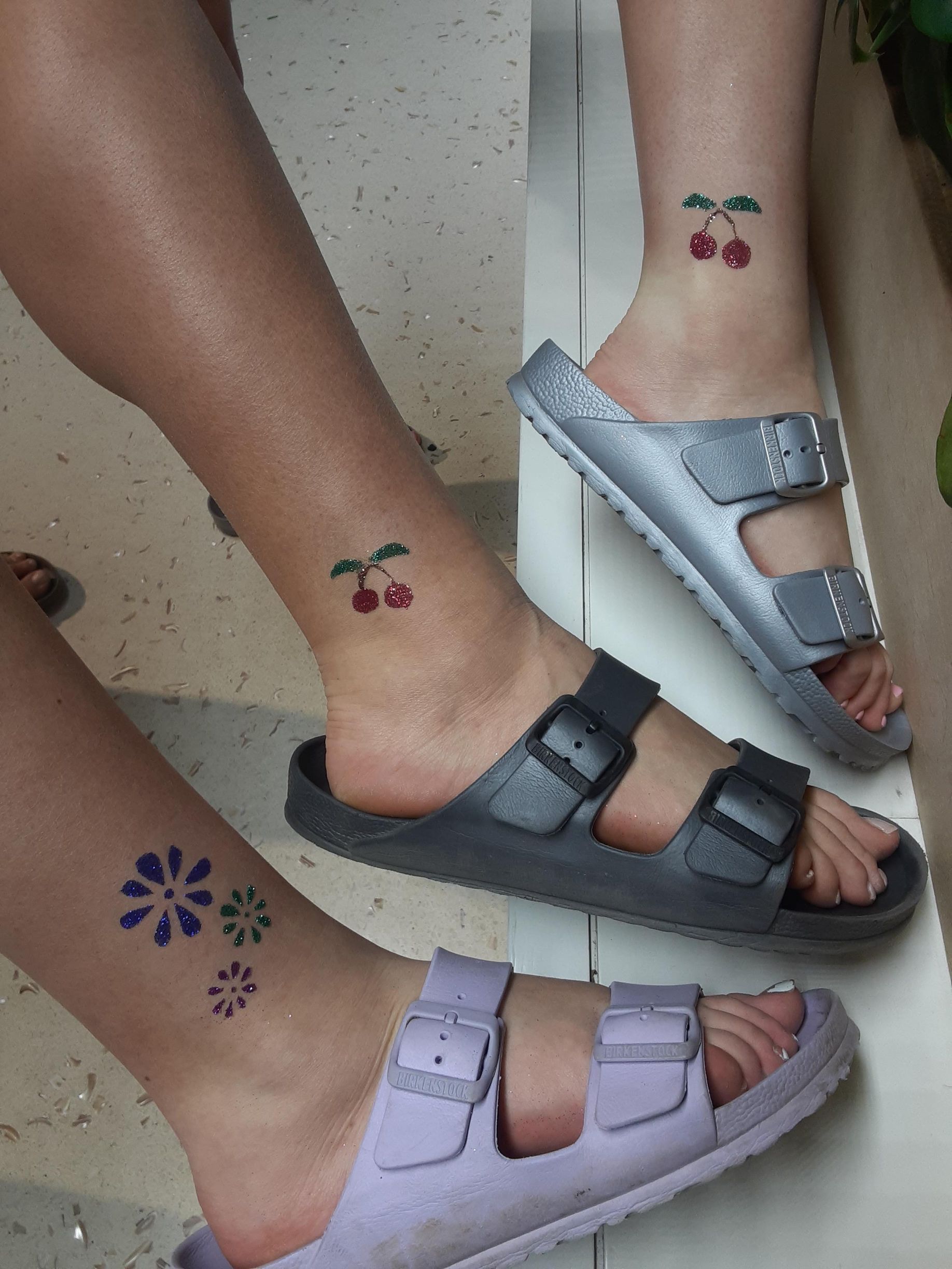 Three people's ankles with matching cherry tattoos and flower tattoos, wearing sandals.