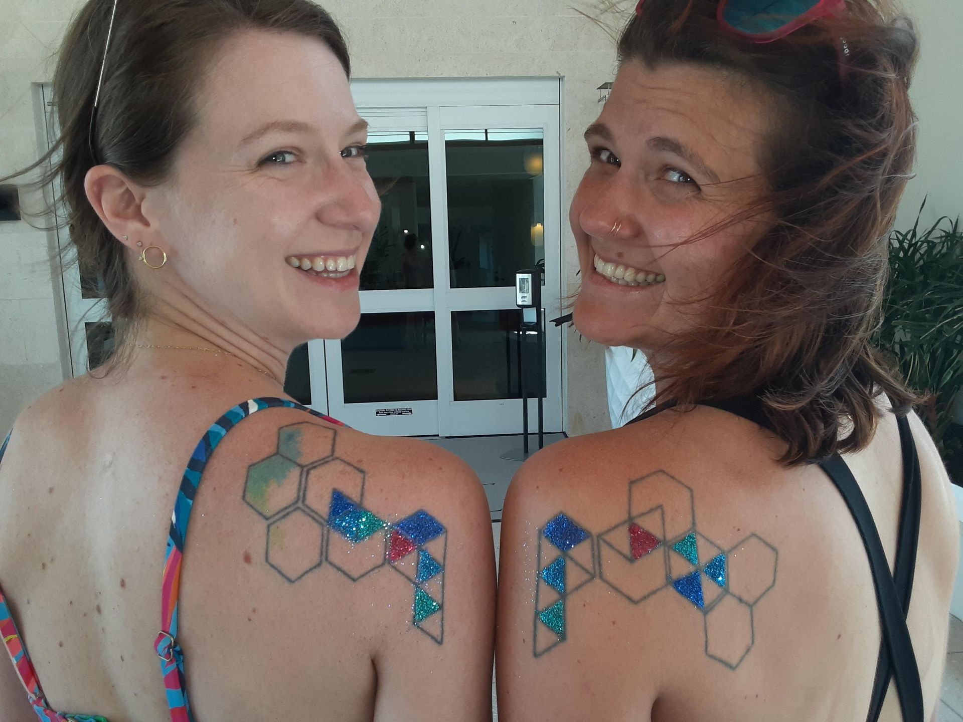 Two women with matching geometric tattoos, smiling, near a building entrance.