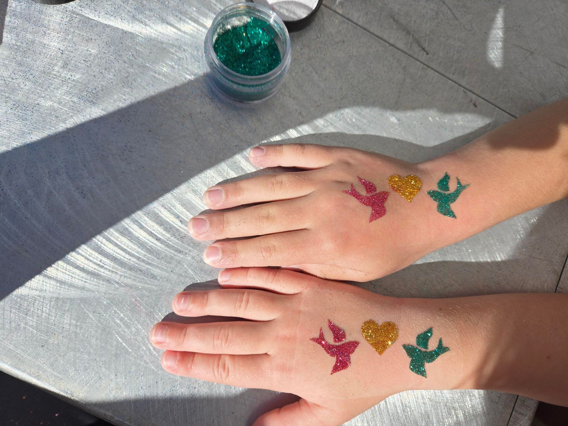 Hands with glitter tattoos: red bird, yellow heart, green bird. Green glitter pot on table.