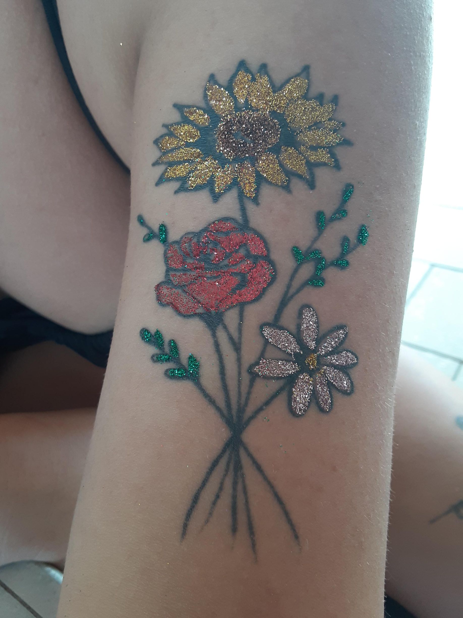 Glittery floral tattoo of a sunflower, rose, and daisy on arm.