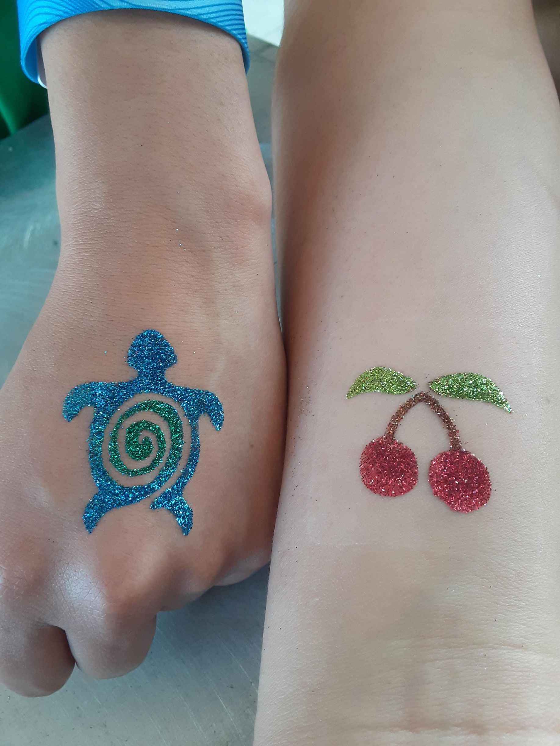 Glitter tattoos a blue turtle on a hand and red cherries on a forearm.
