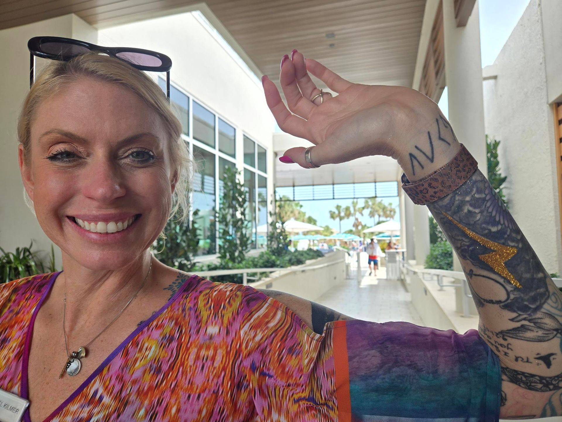 Woman smiles, raises arm to show tattooed sleeve; sunny hallway with glass windows and outdoor view.