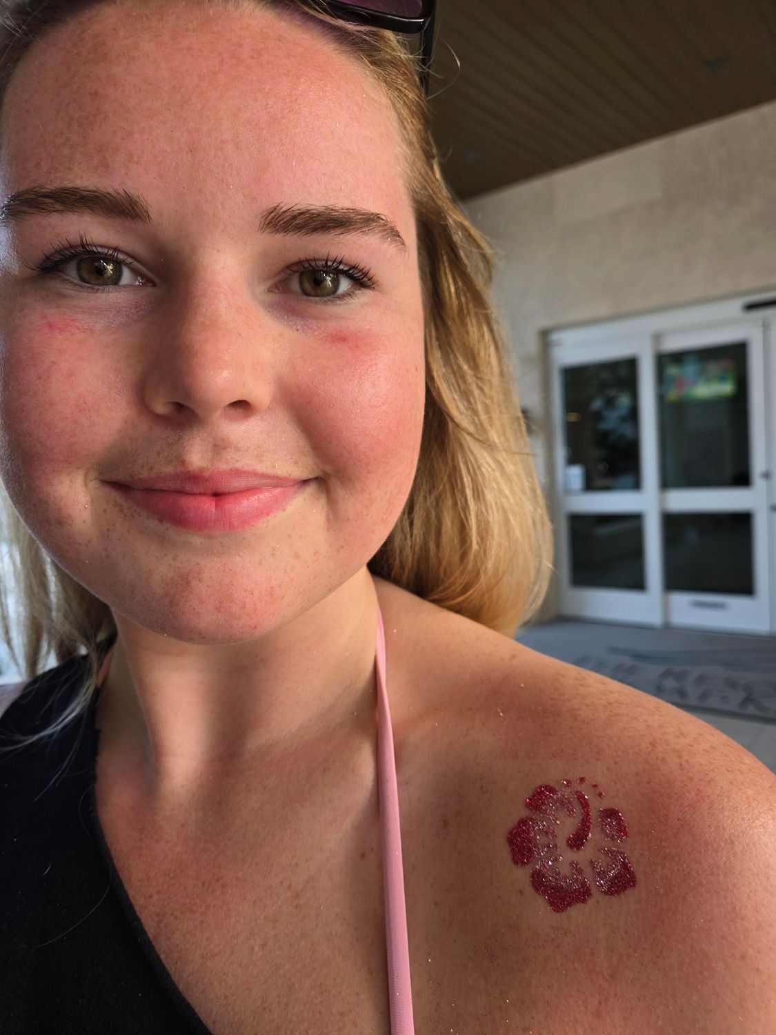 Smiling woman with a temporary flower tattoo on her shoulder. Outdoors, near a building with glass doors.