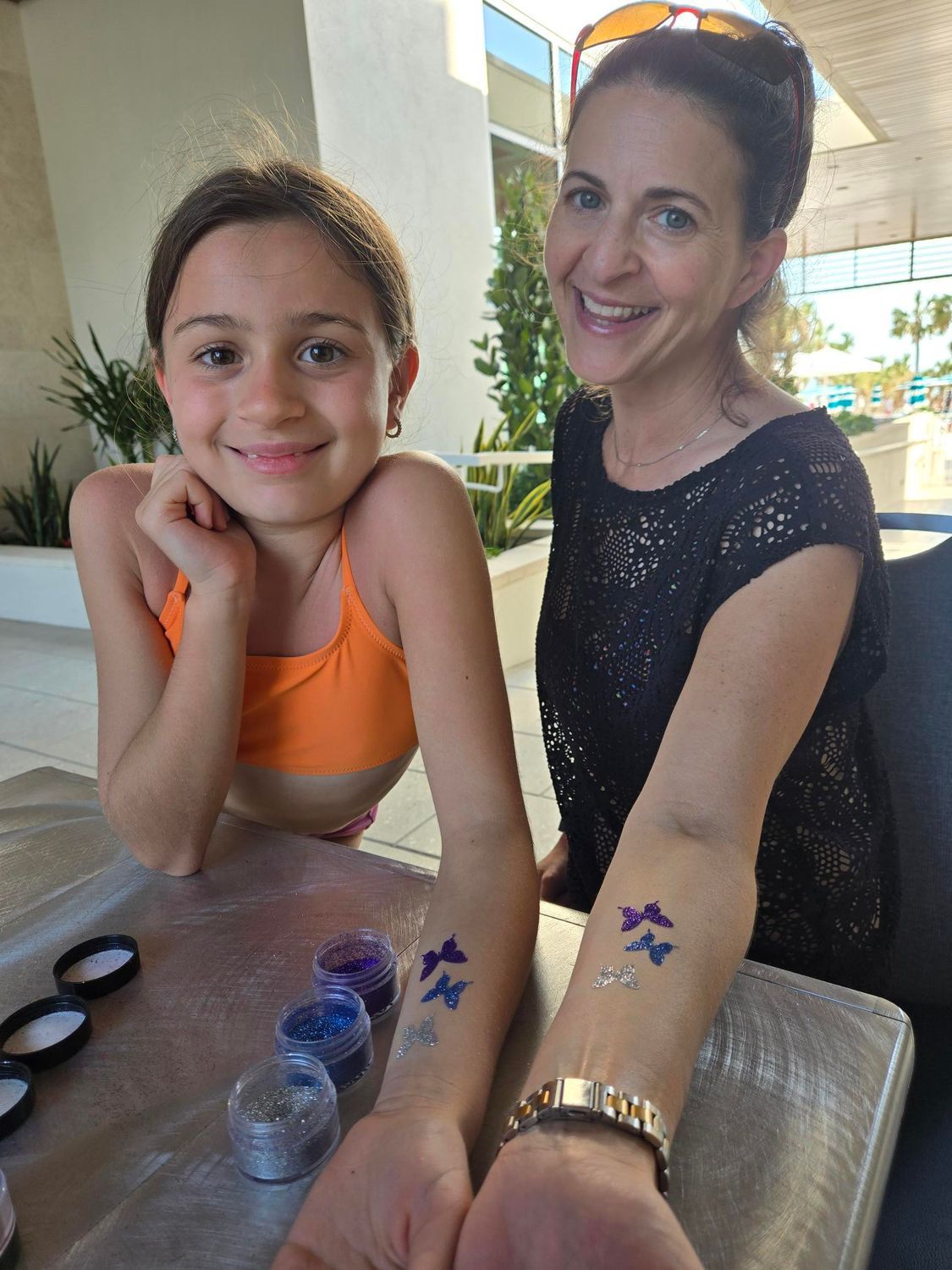 Woman and girl with glitter tattoos, smiling, outdoors.  Purple and silver butterflies on arms.
