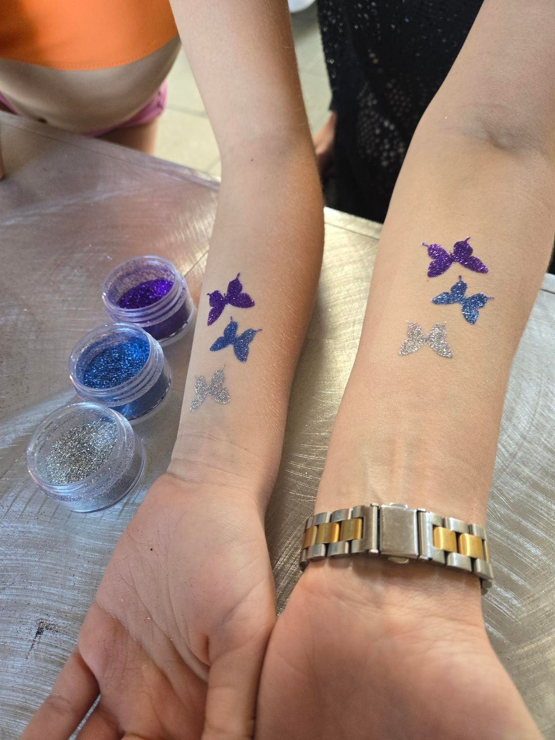 Two forearms with glitter butterfly tattoos in purple, blue and silver, next to jars of glitter.
