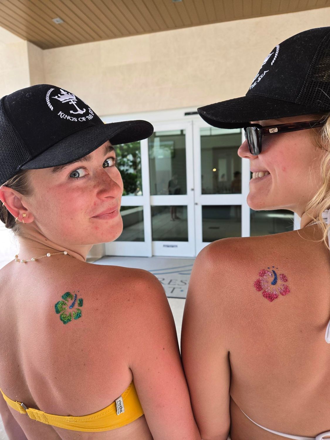 Two women with matching shoulder tattoos, smiling, wearing hats and bikinis.