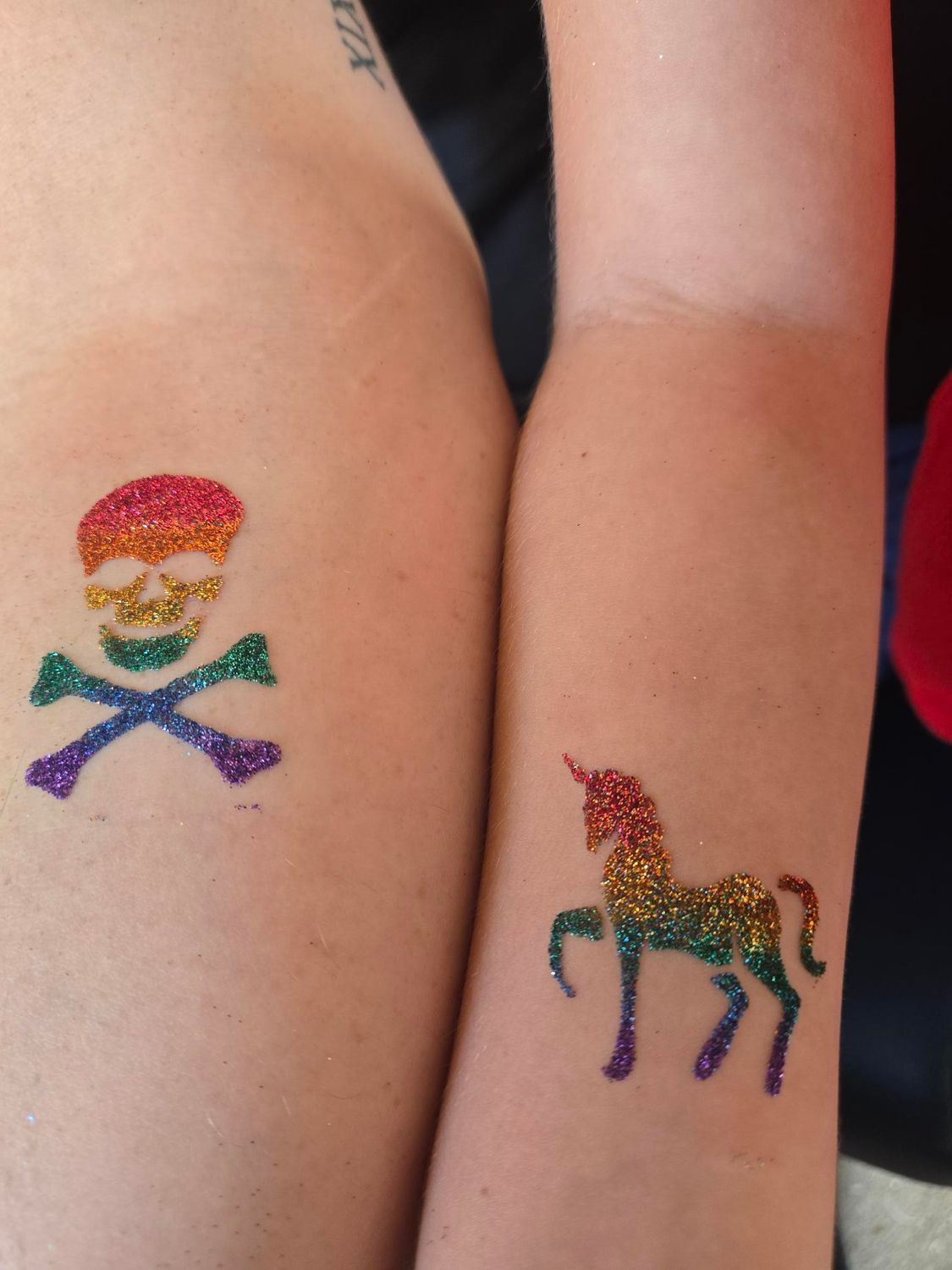Two arms with glitter tattoos: a rainbow skull and crossbones, a rainbow unicorn.