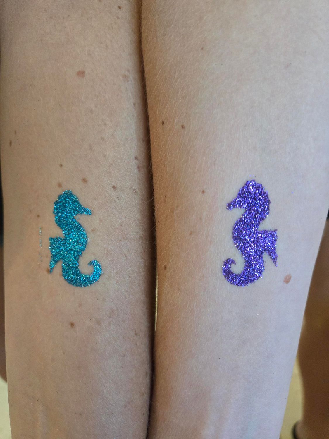 Two arms with matching glitter seahorse temporary tattoos, one teal, one purple.
