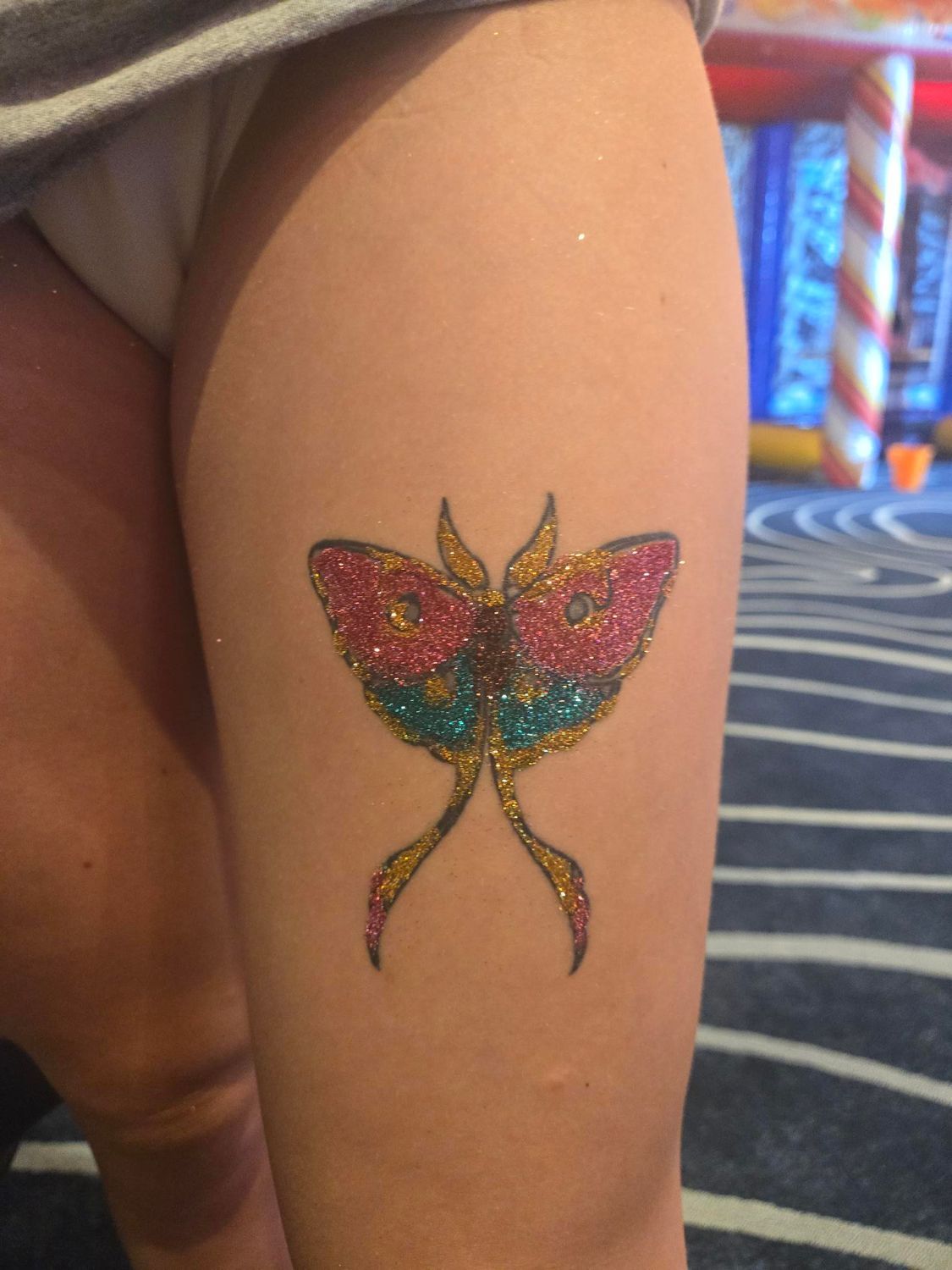 Glittery, colorful moth tattoo on thigh, pink, teal, gold wings; long pink streamers.