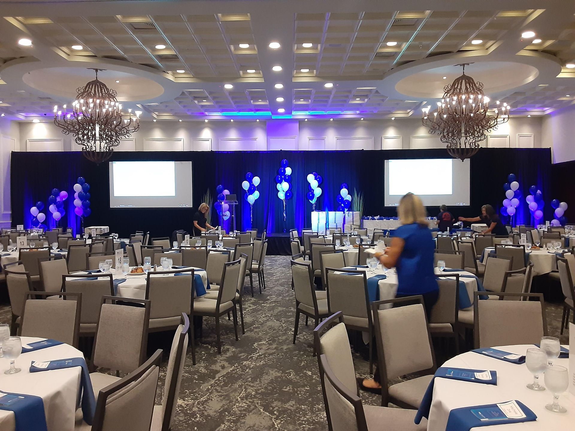 A woman in a blue shirt is standing in a large room filled with tables and chairs.