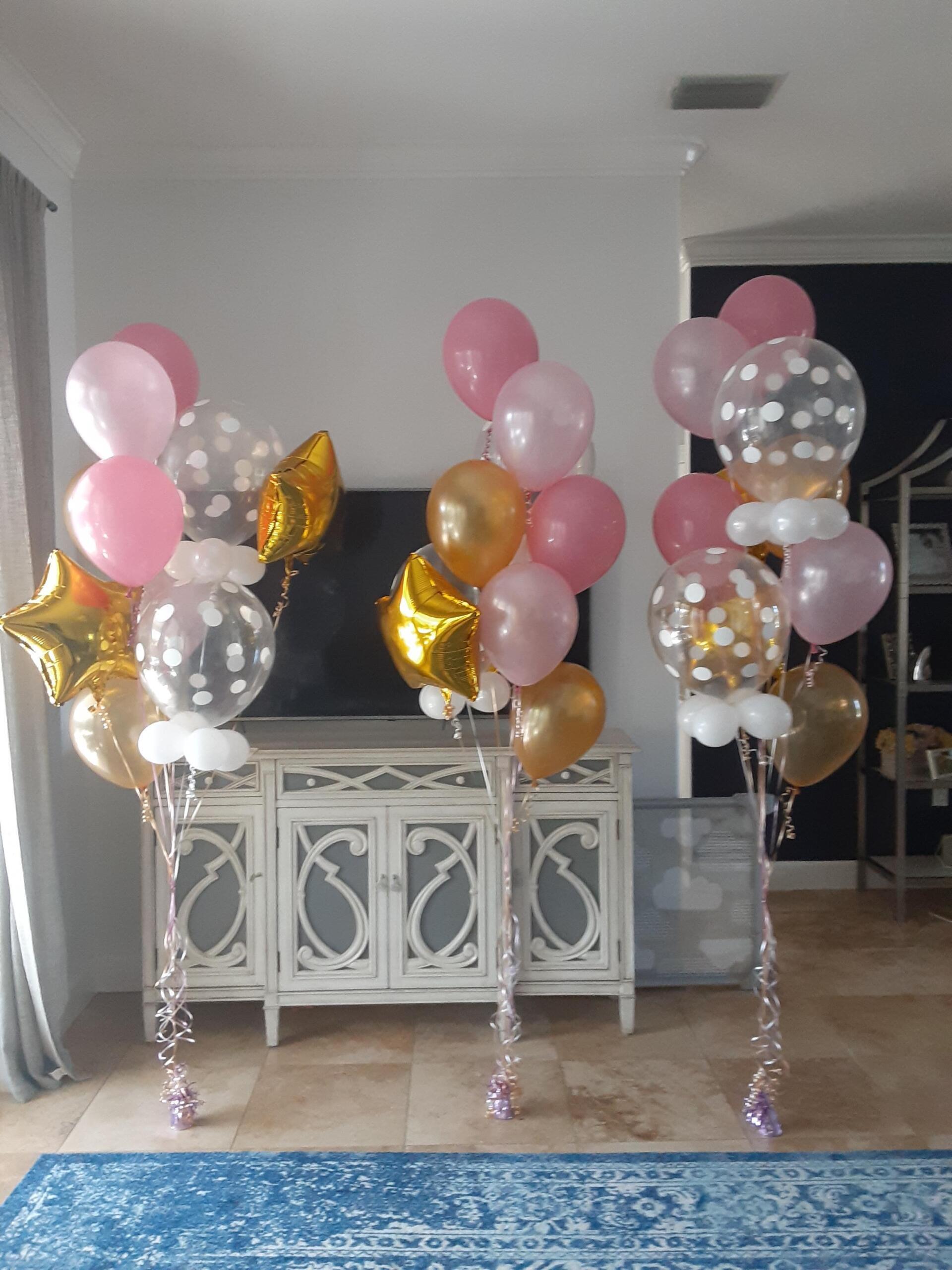 Three bunches of pink and gold balloons are sitting in a living room.