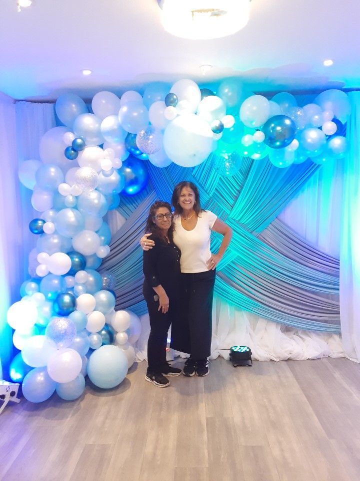 Two women are standing in front of a blue and white balloon arch.